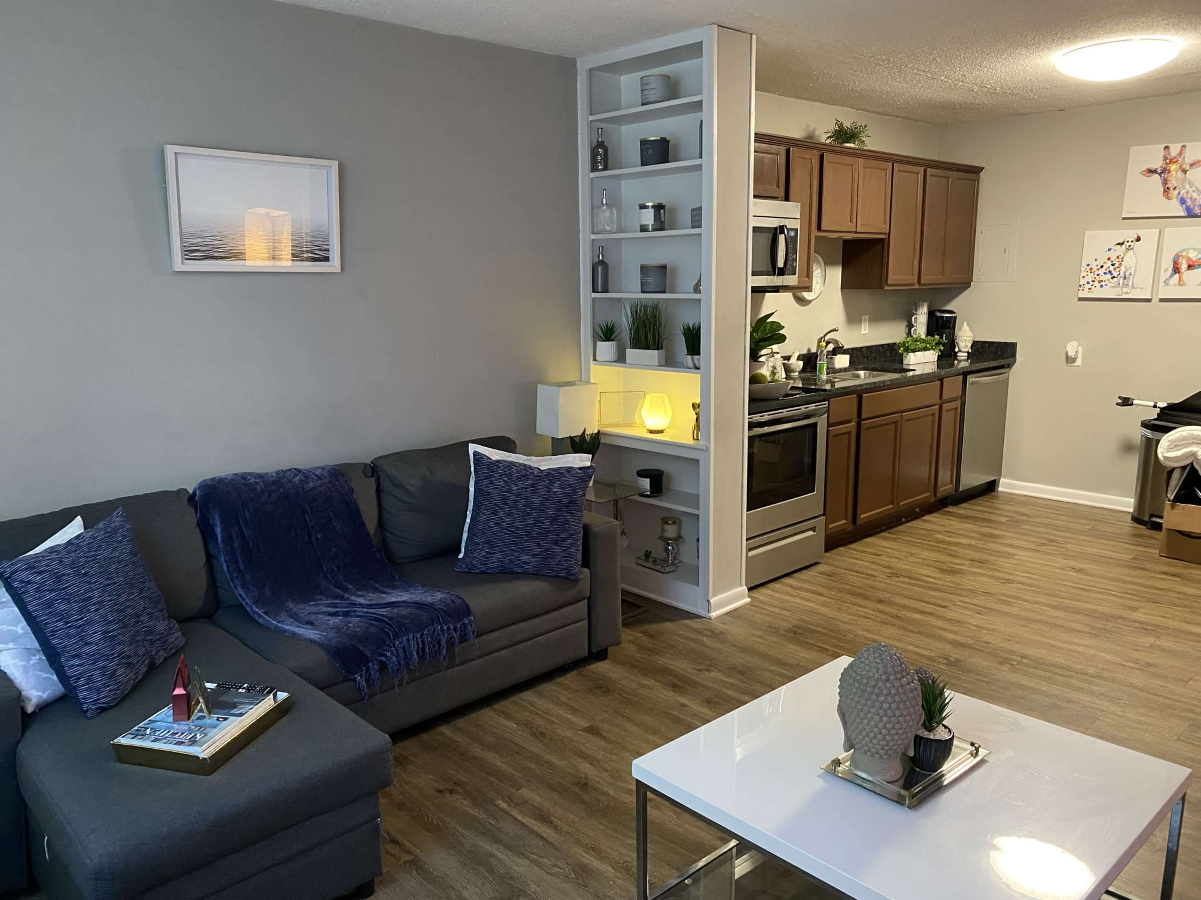 The image shows a modern living room with a gray sofa, a white coffee table, a kitchen in the background, and decorative shelving featuring plants and a light source.