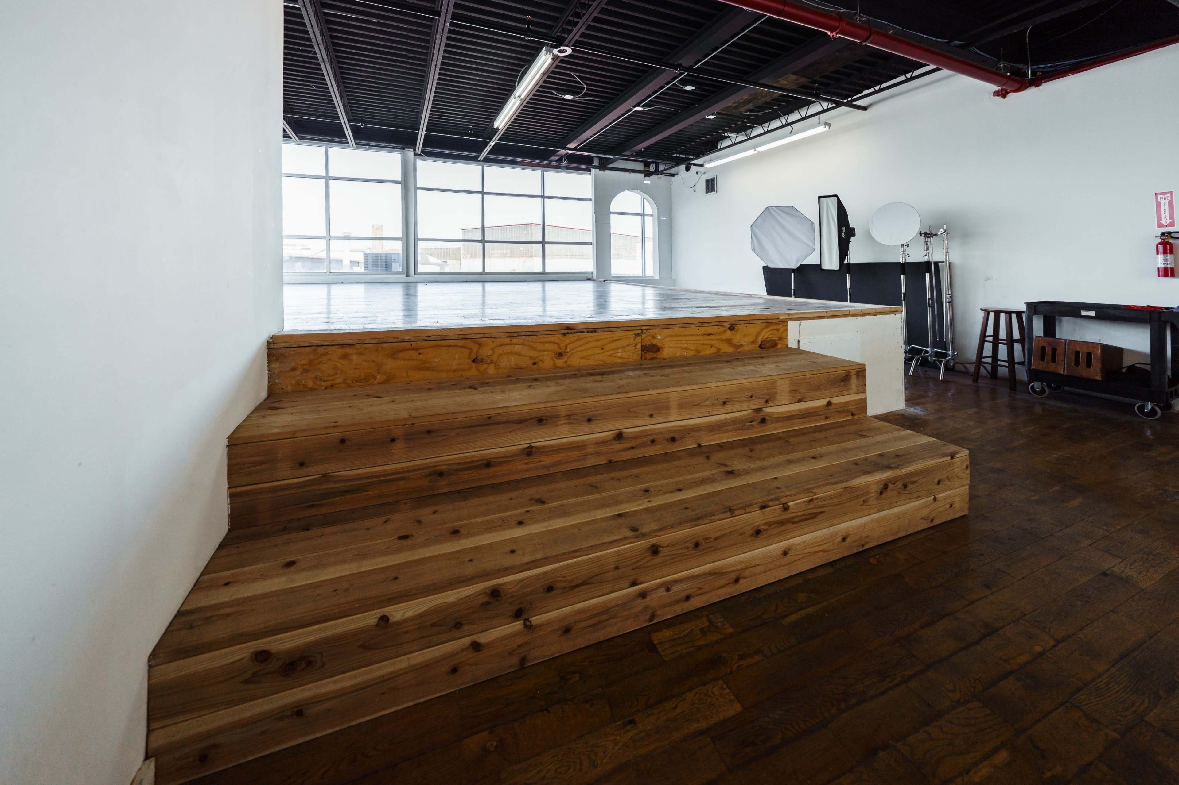 A spacious studio with wooden steps leading up to a raised platform, surrounded by large windows and photography equipment.