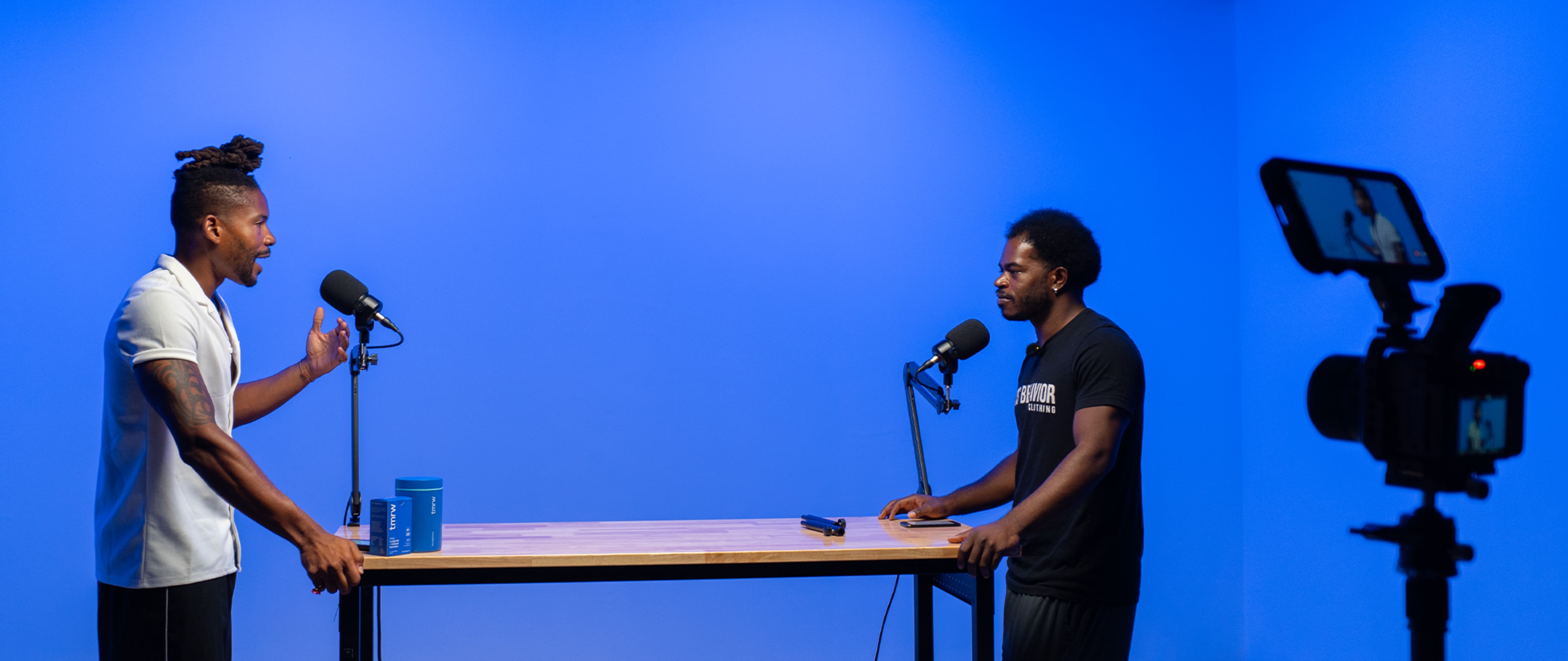 Two men are engaged in conversation at a wooden table with microphones, against a blue backdrop, while a camera captures the scene nearby.