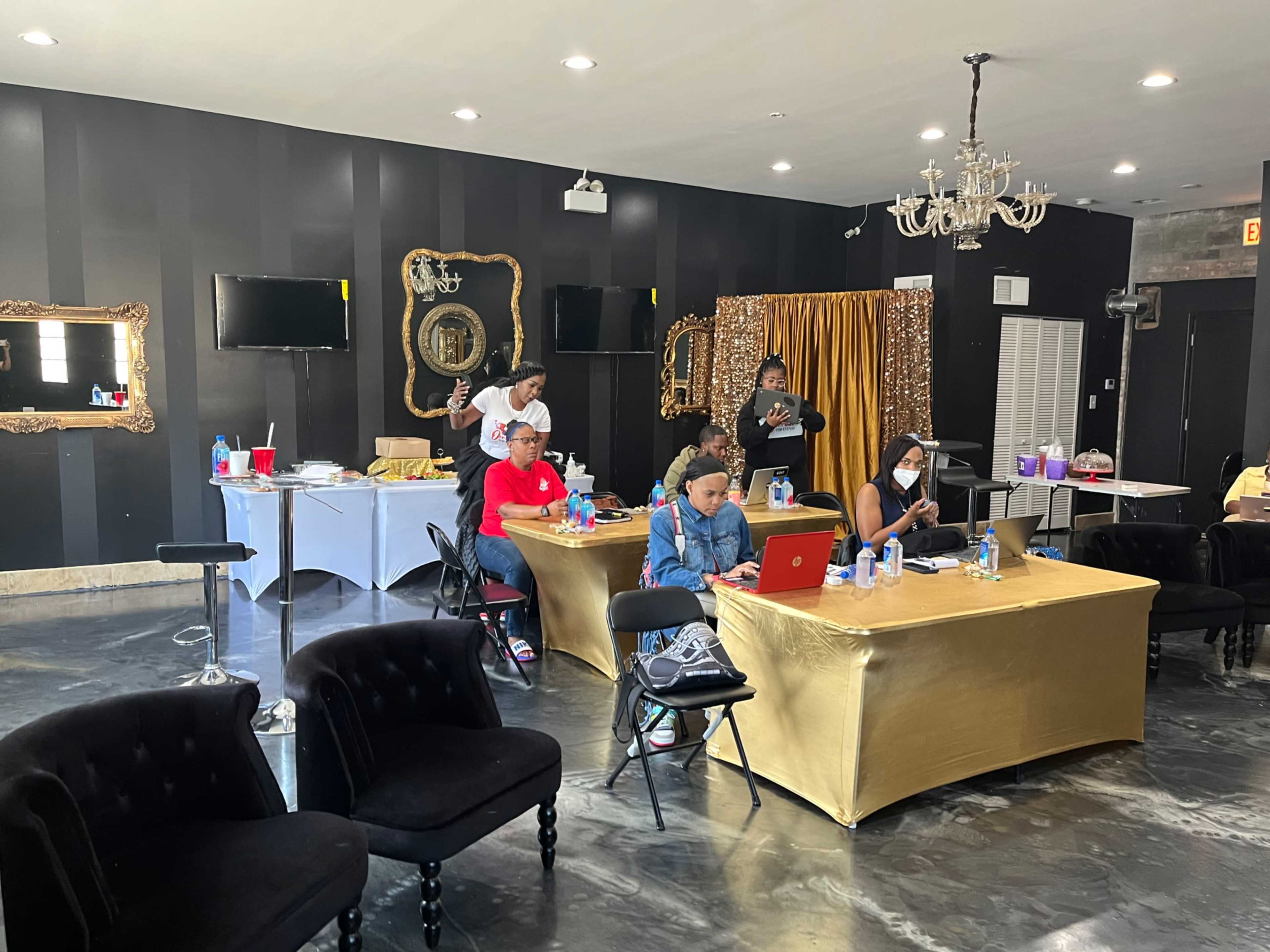 A group of people is gathered in a room with black walls and elegant decorations, working on laptops while some prepare food at a table in the background.
