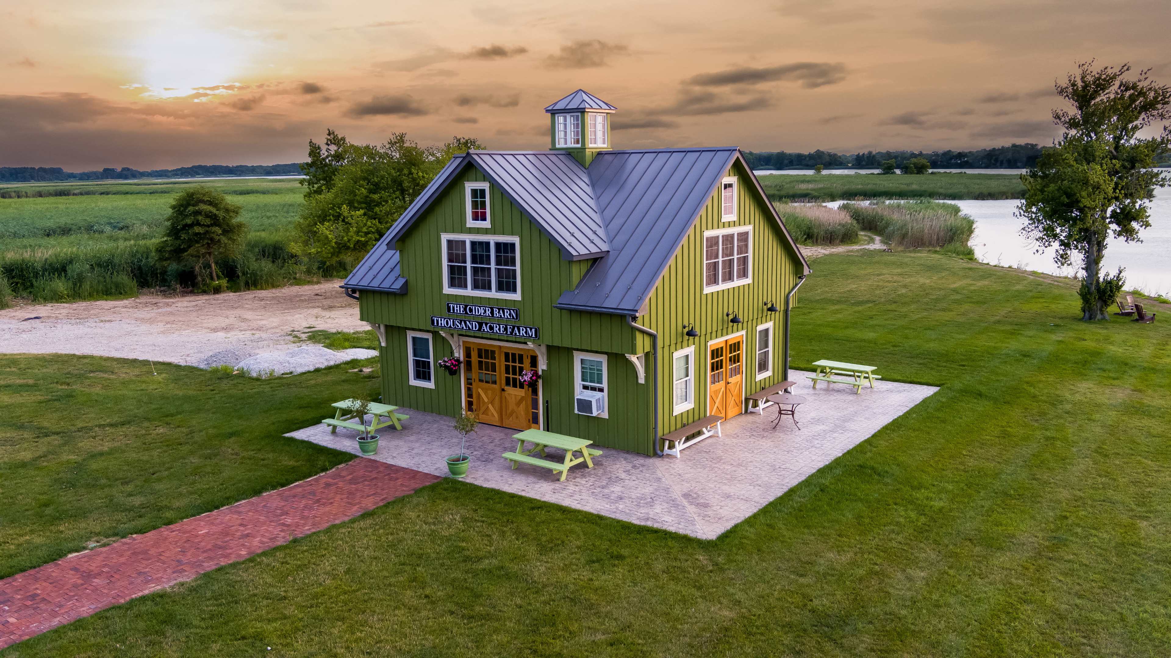 A green barn-style building with a metal roof stands near a calm body of water, surrounded by grassy areas and picnic tables.