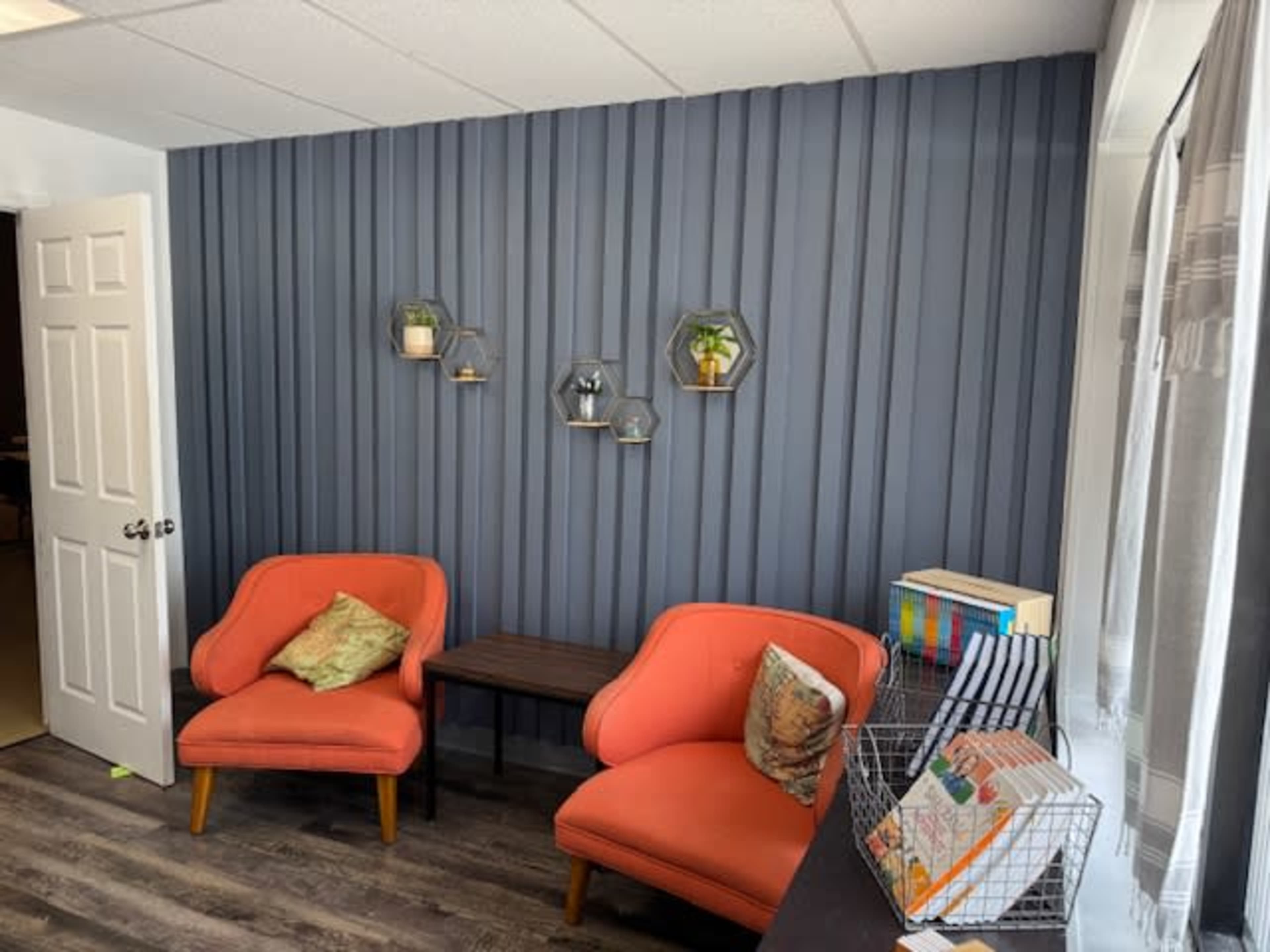 The image shows a small, bright room featuring two orange chairs, a wooden table, and decorative wall-mounted shelves with plants.