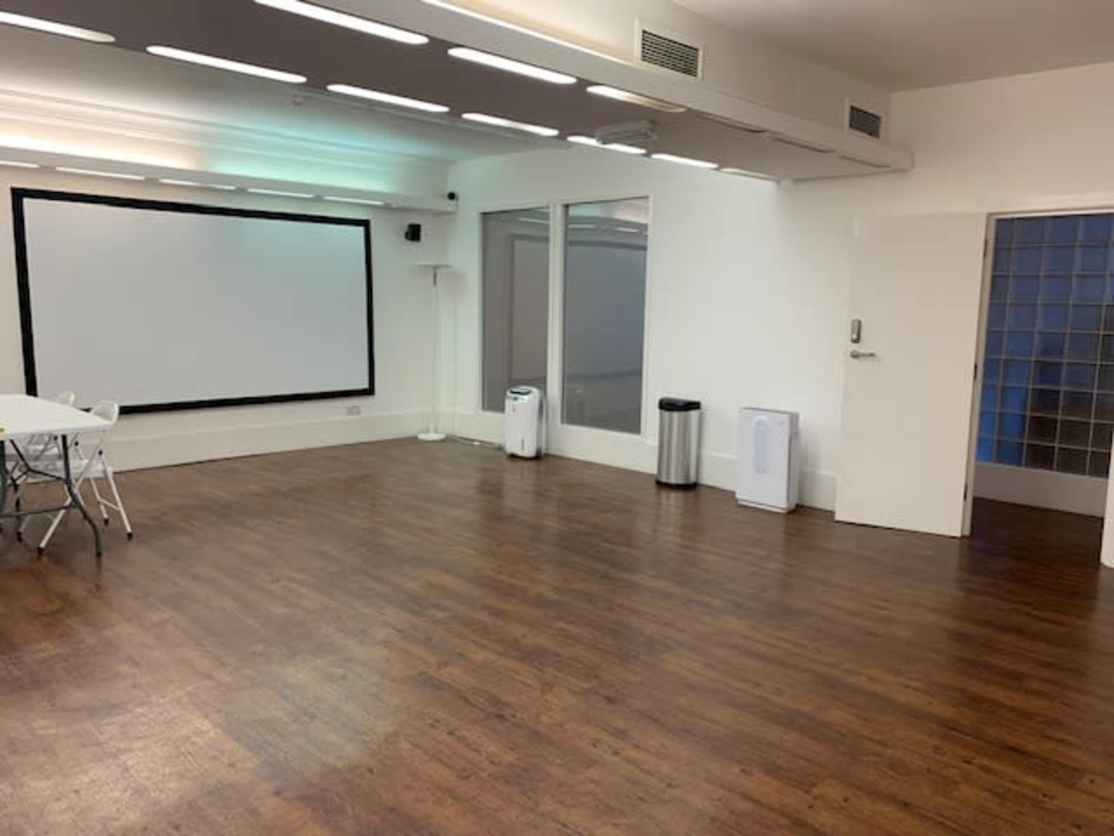 An empty room features a large projector screen, a table with chairs, a trash can, and a door leading to a glass-block wall.