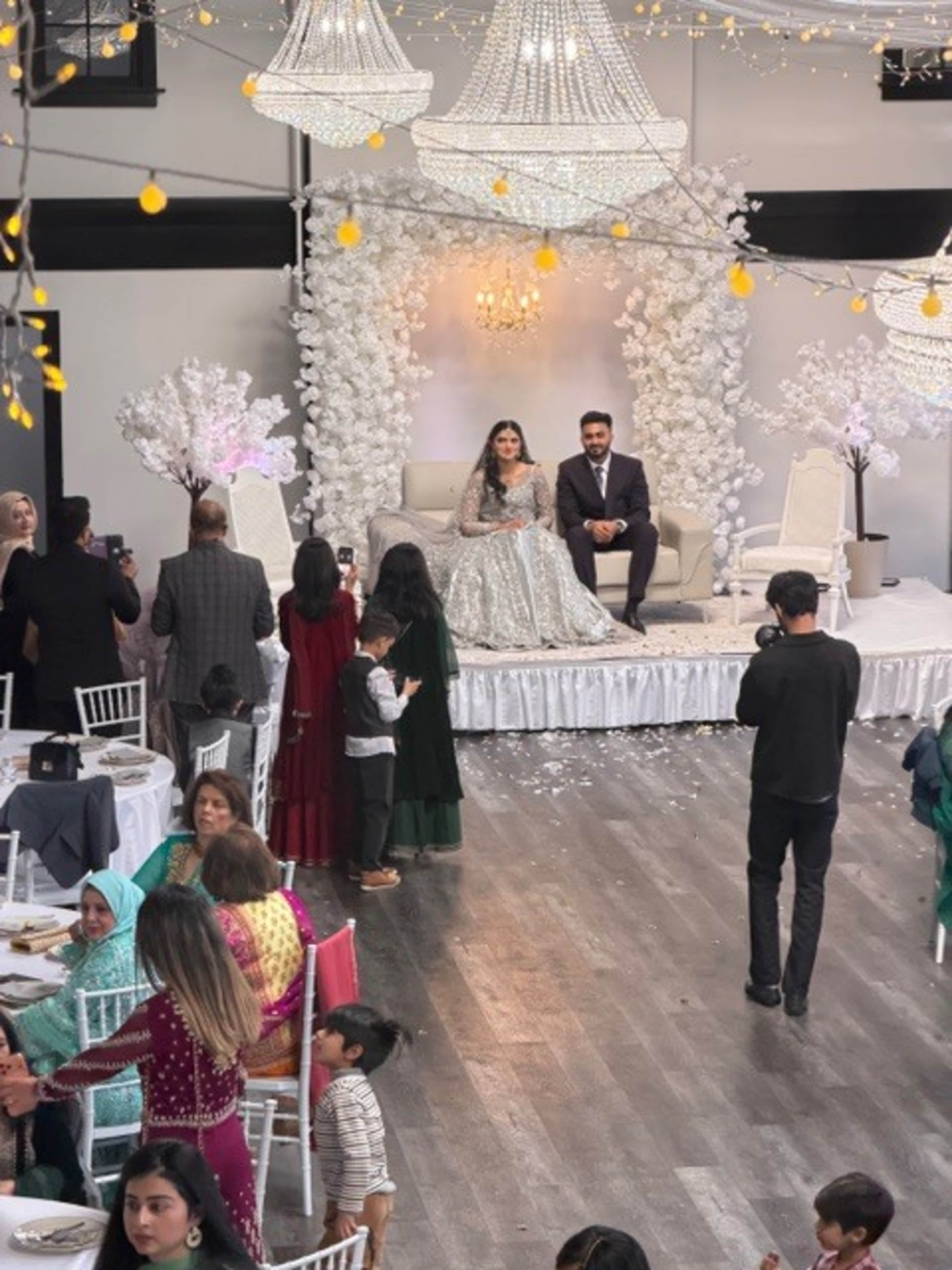 A bride and groom sit on an ornate stage decorated with white floral arrangements and chandeliers while guests enjoy a meal in a festive reception hall.