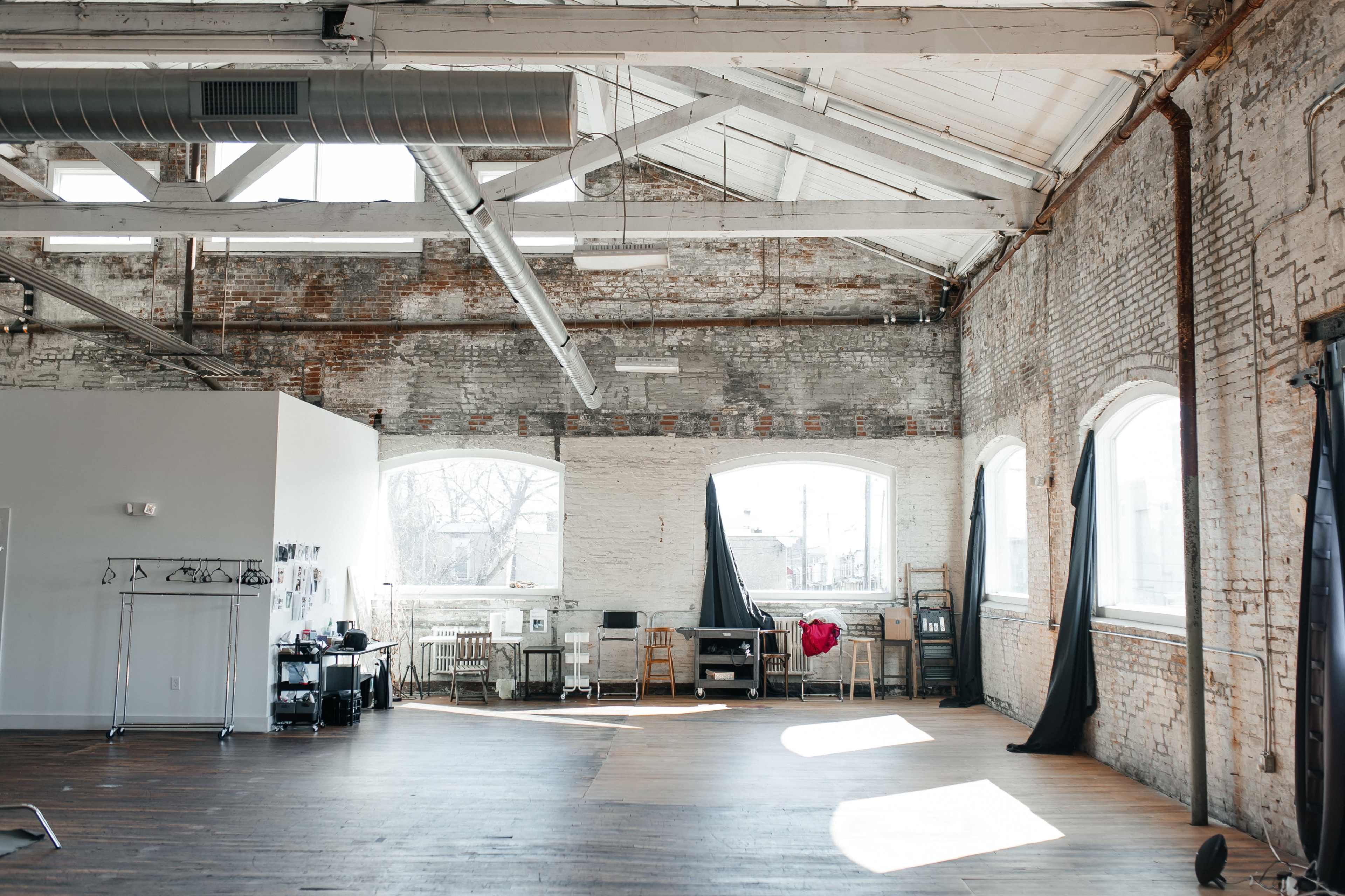 An industrial-style interior space with large windows, exposed brick walls, and wooden flooring, featuring scattered furniture and a black curtain in the corner.