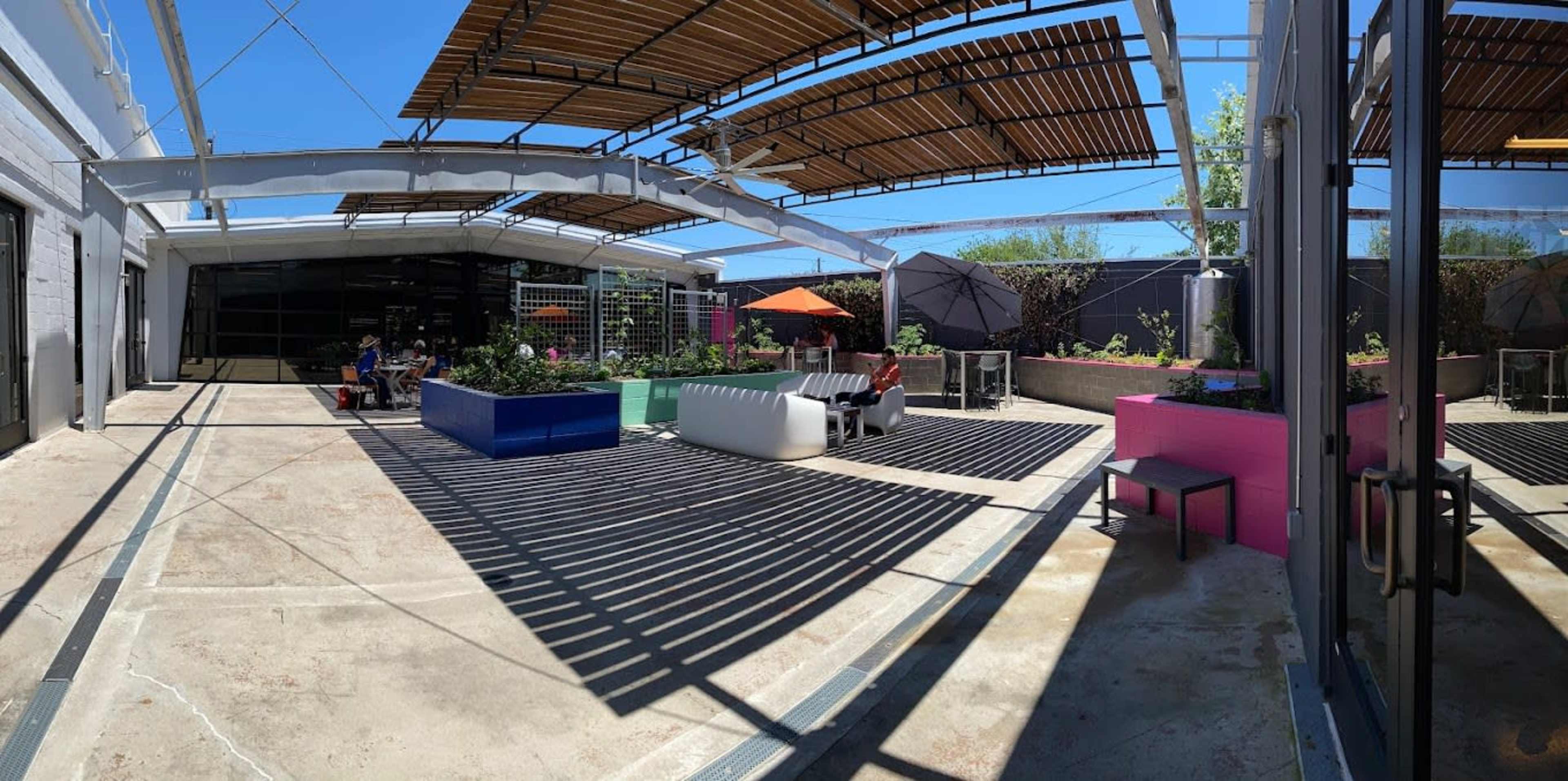 The image shows an open courtyard with seating areas, colorful furniture, and shaded spaces under a large roof structure.