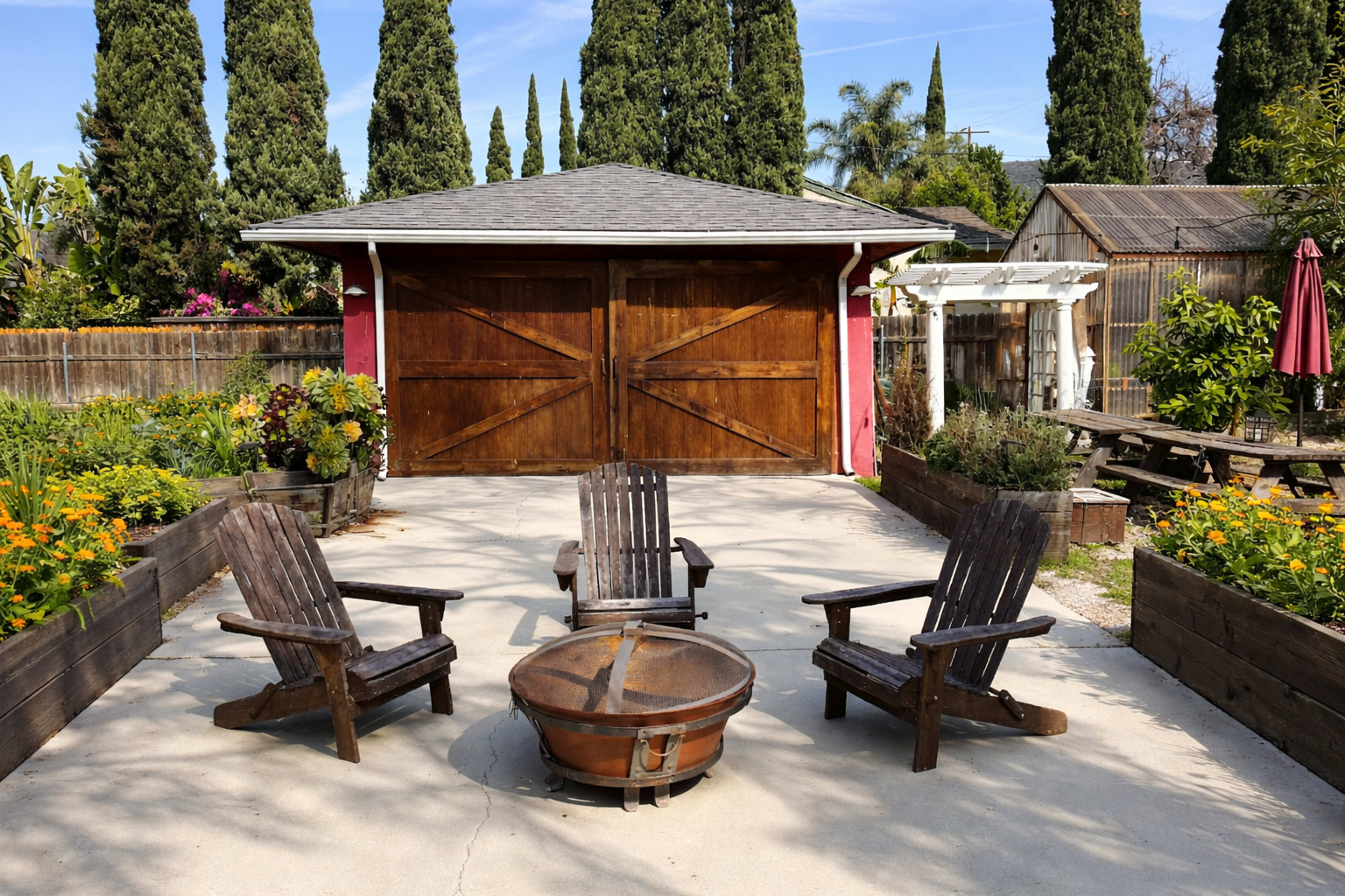 The image shows a backyard scene with four wooden Adirondack chairs arranged around a circular fire pit, in front of a wooden garage with double doors, surrounded by greenery and colorful flowers.