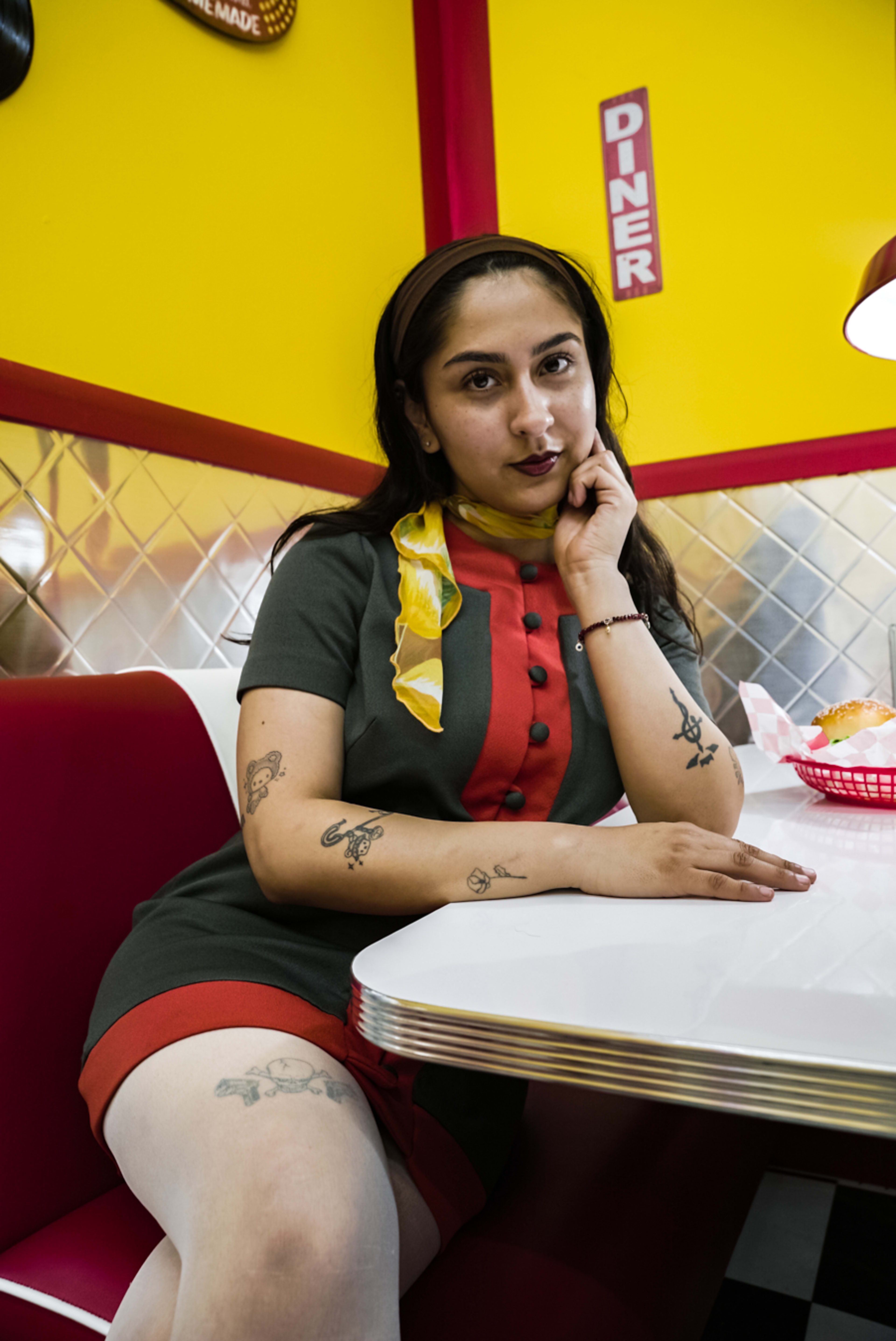 A woman with tattoos sits at a diner booth, resting her chin on her hand, wearing a retro-style dress and a colorful scarf.