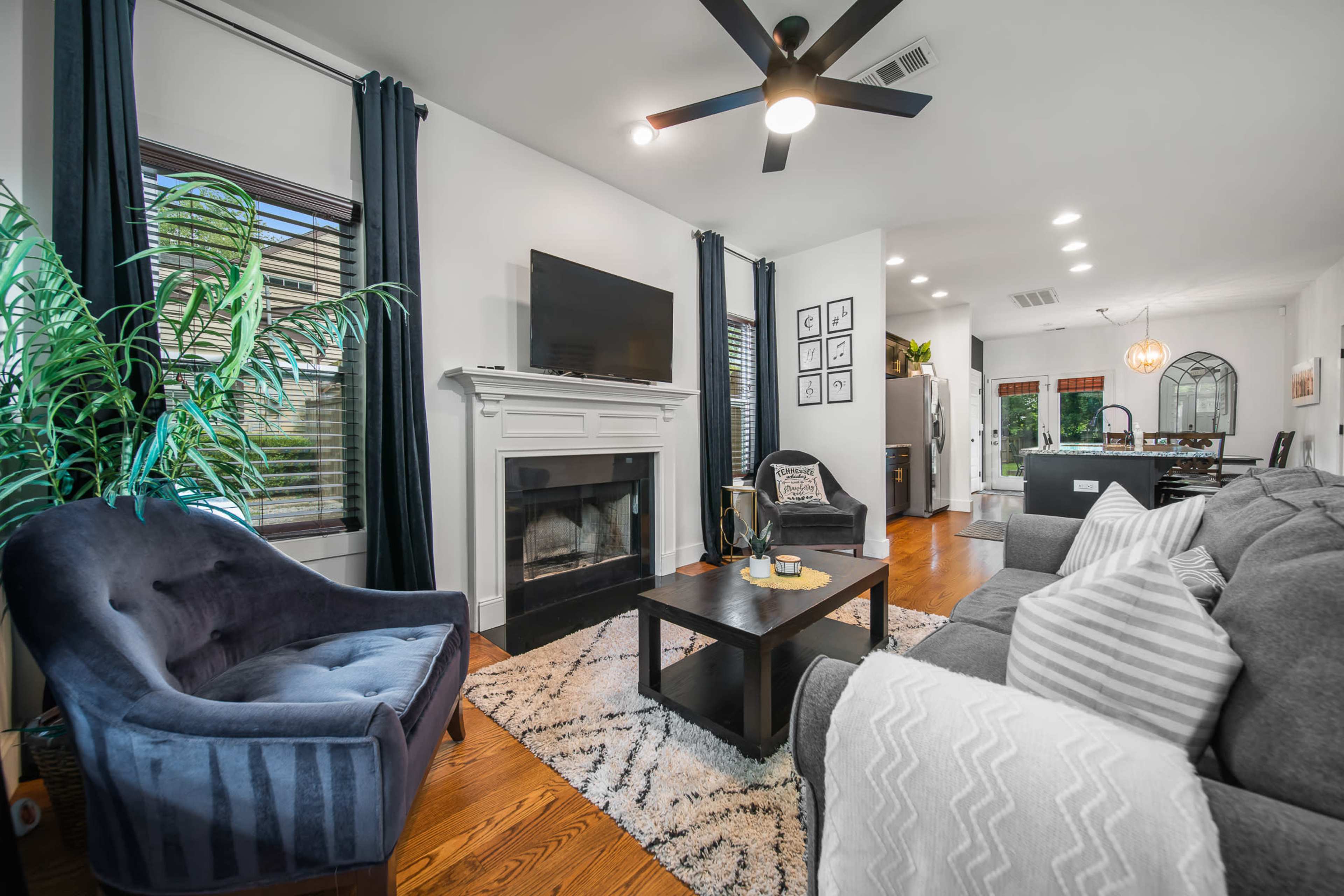 A cozy living room features a fireplace, a dark blue armchair, gray sofas, and light hardwood flooring, with large windows dressed in dark curtains.