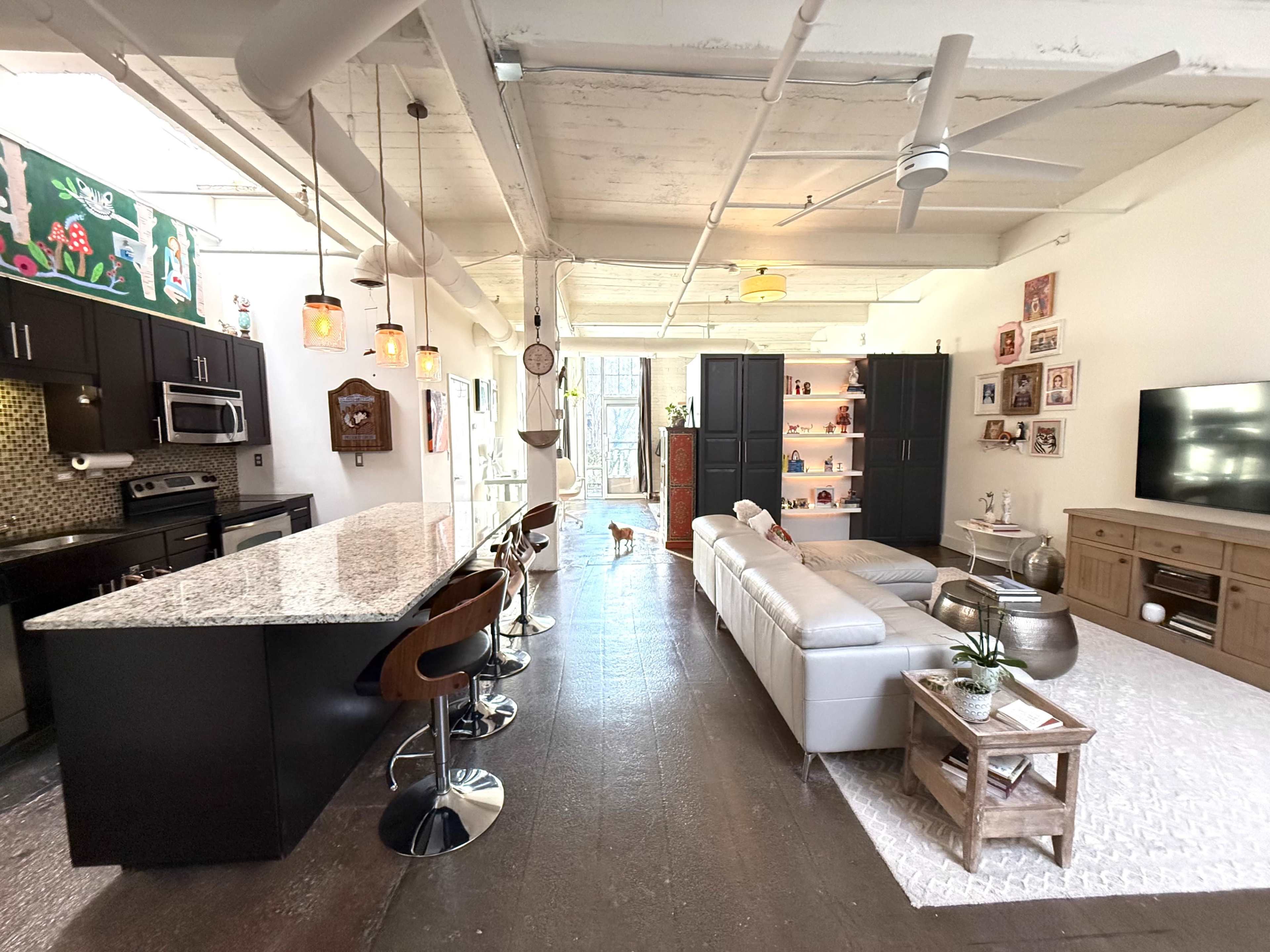 The image shows a modern open-concept living space with a kitchen island, seating area, and large windows allowing natural light to enter.