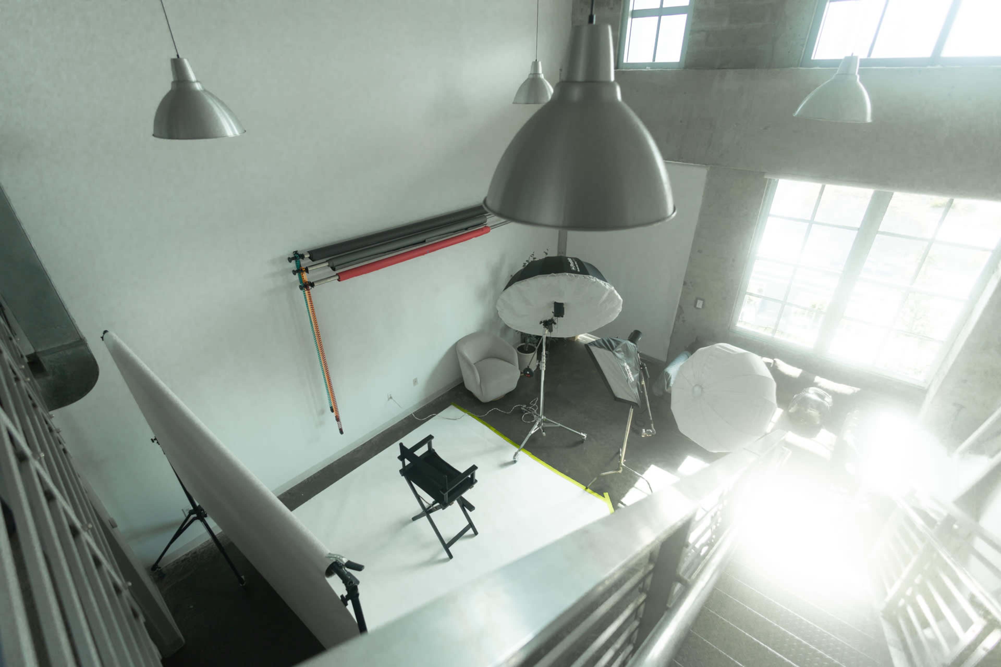 A photography studio features a softbox lighting setup, a director's chair, and a backdrop against a large window with natural light flooding in.