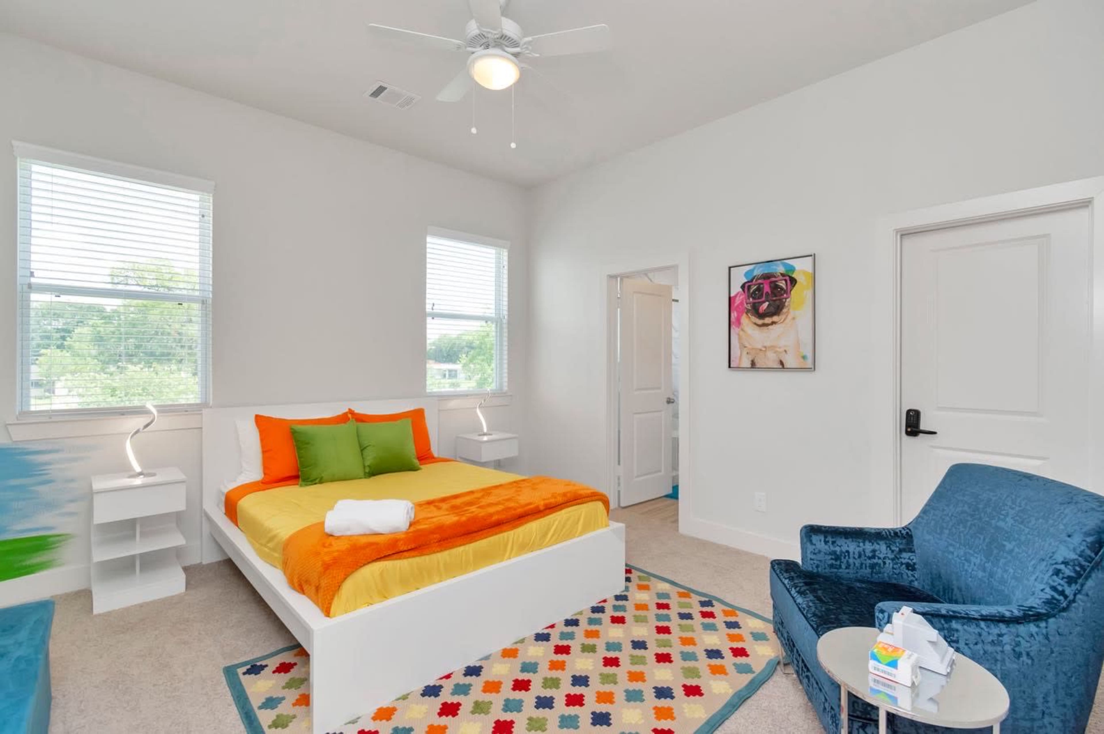 A brightly decorated bedroom with a bed covered in orange and green bedding, a blue armchair, and a colorful rug on the carpeted floor.