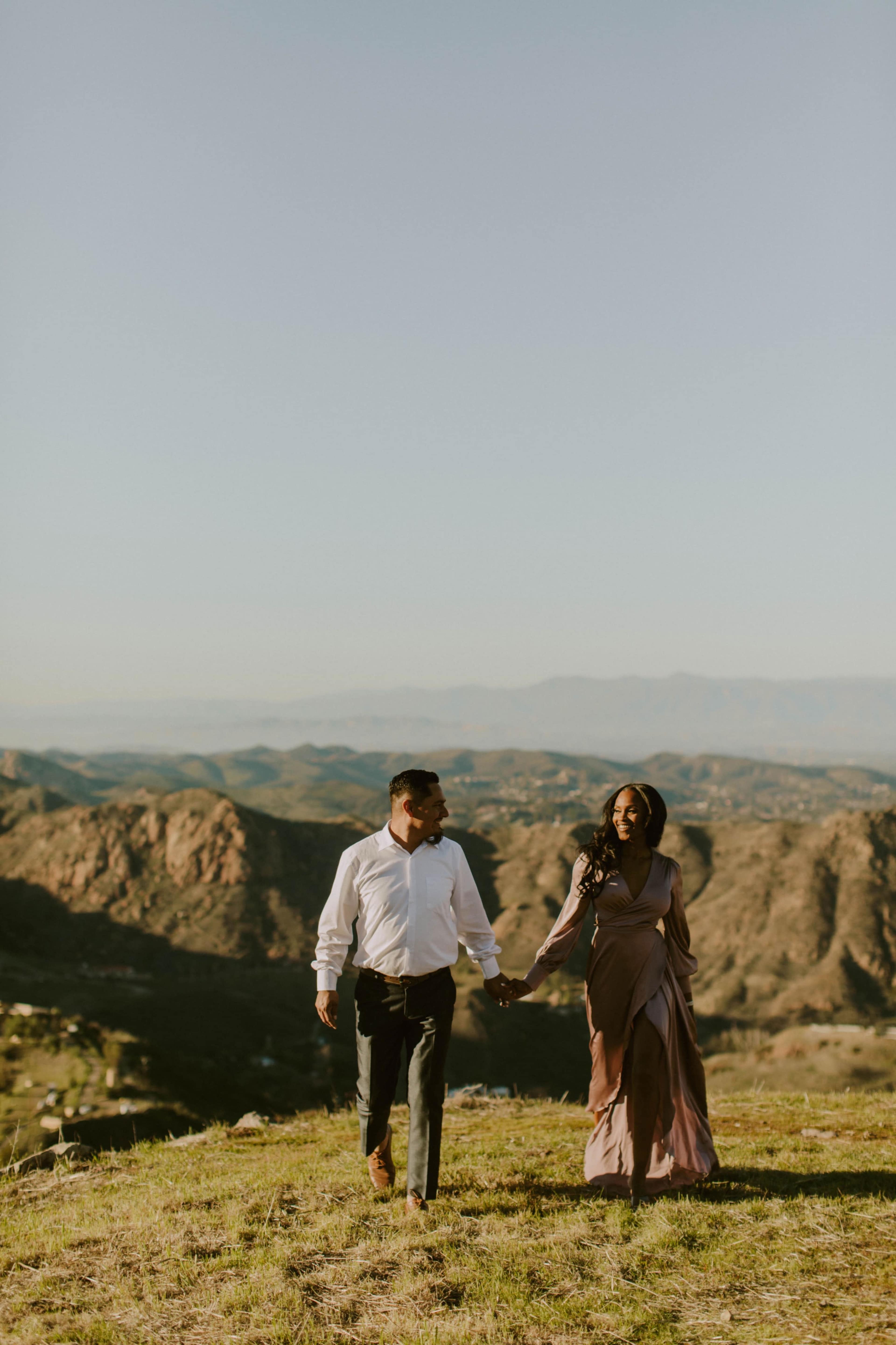A couple walks hand in hand on a grassy hillside with mountains in the background.