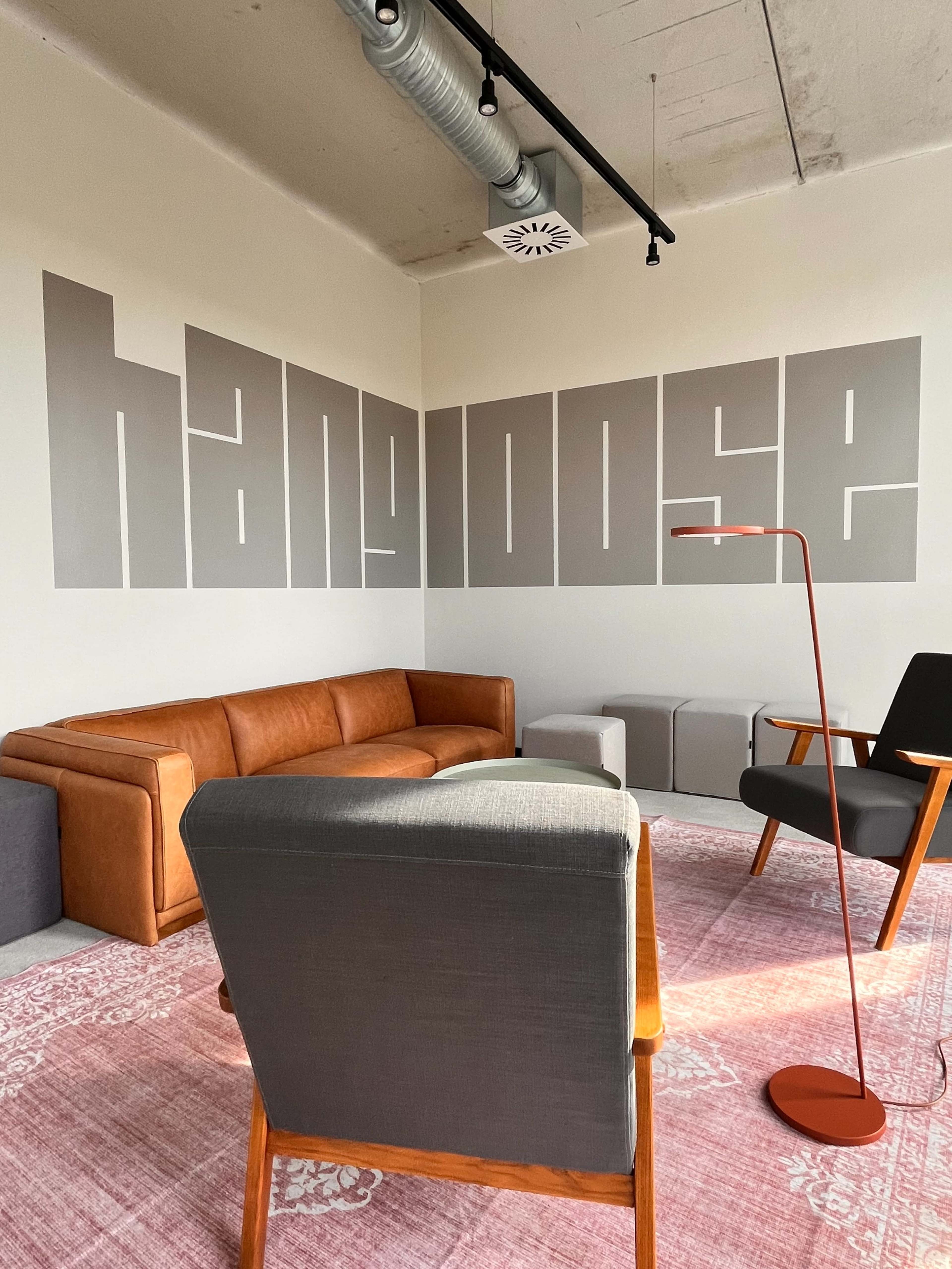 The image shows a modern room featuring a brown leather sofa, gray upholstered chairs, a pink patterned rug, and a large wall with the word "hangloose" displayed in bold letters.