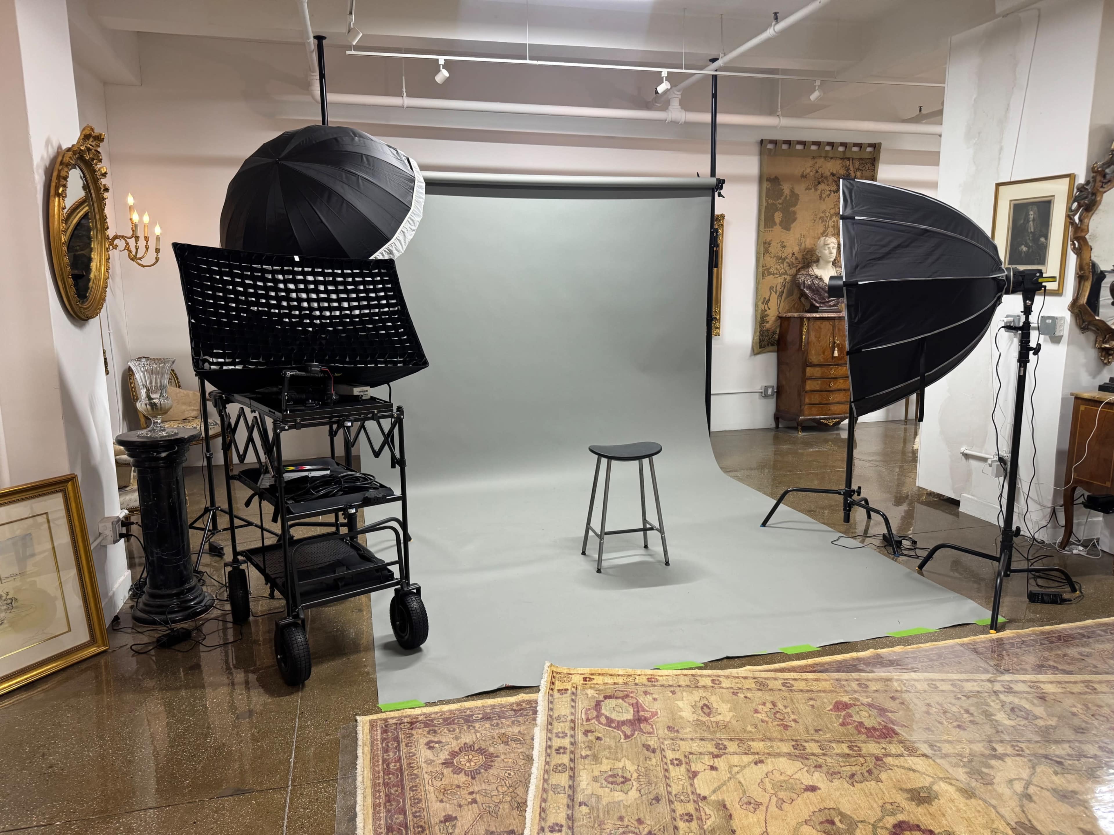 The image shows a photography studio setup with a gray backdrop, a stool in the center, and lighting equipment on either side.