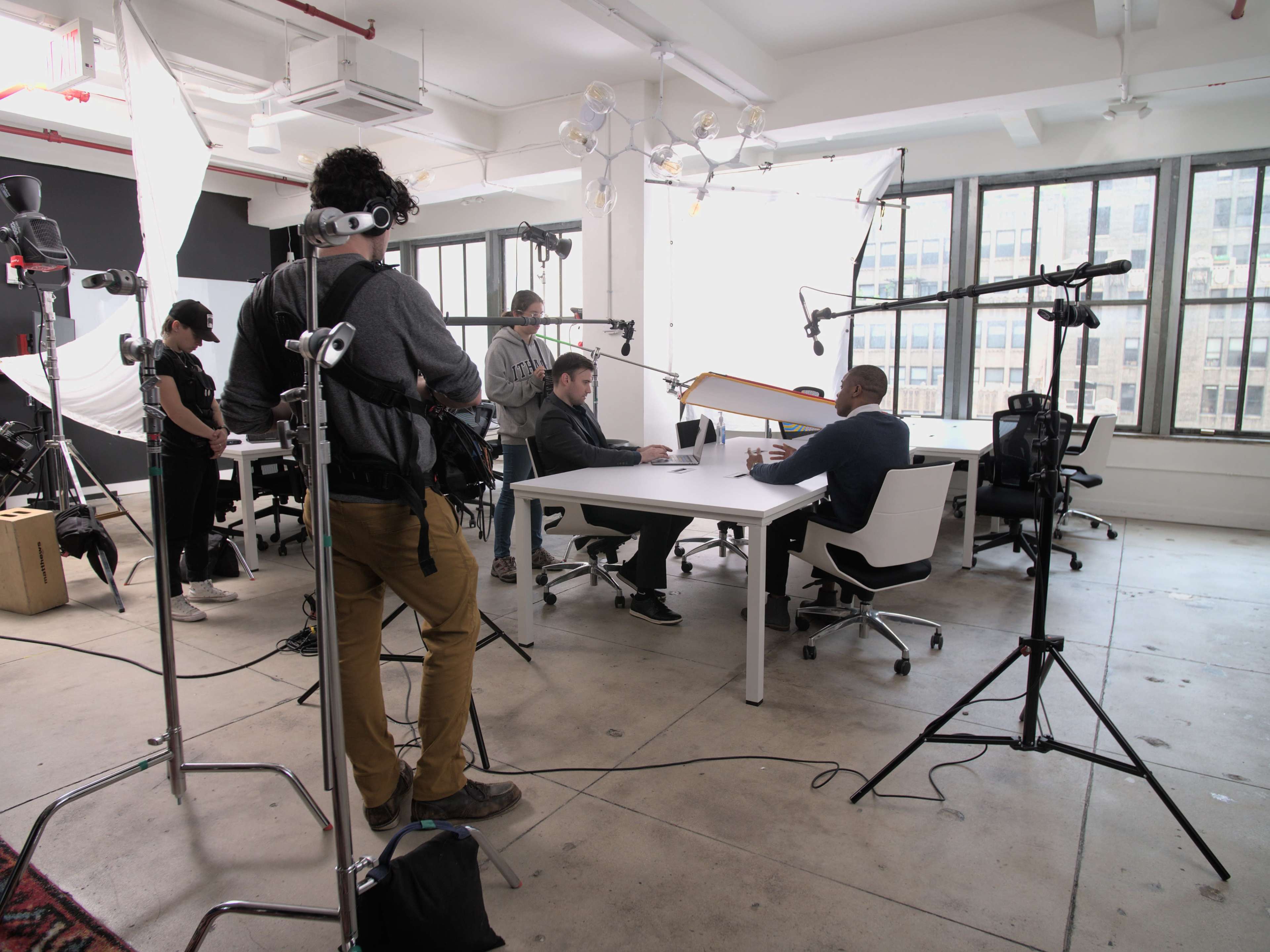 A film crew sets up equipment around a table where two people are engaged in a discussion in a well-lit office space.