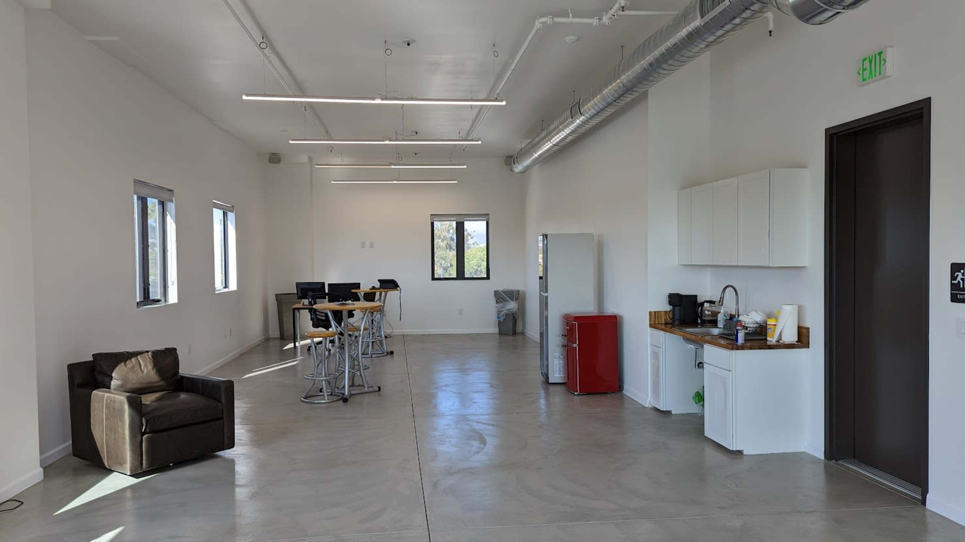 The image shows a spacious interior area with modern furnishings, including a kitchen counter, appliances, seating, and several tables.