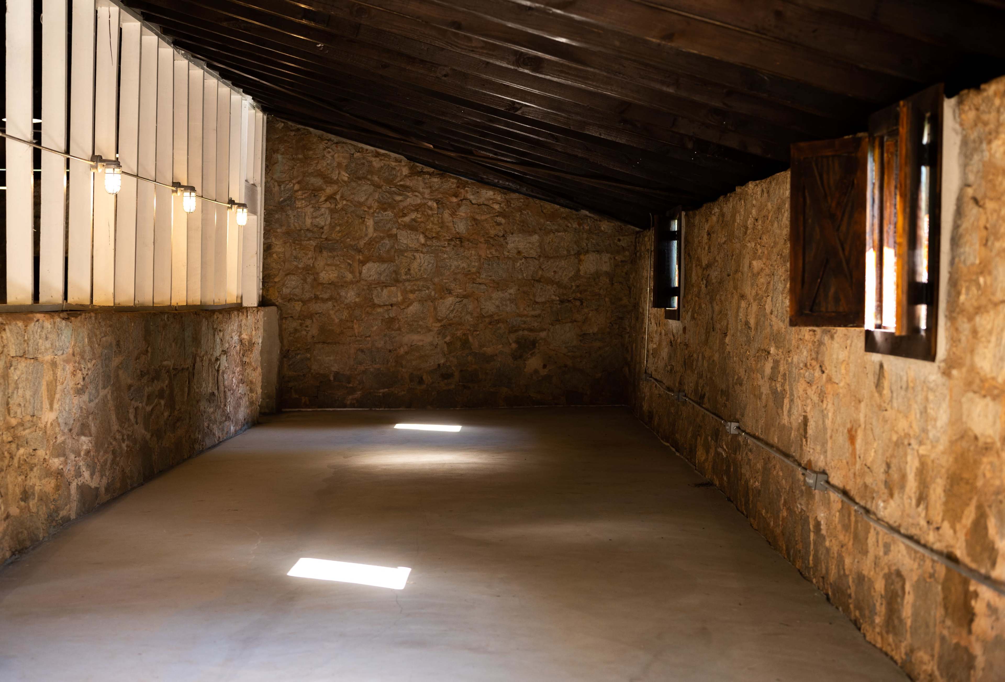 The image shows a stone-walled room with a wooden ceiling, featuring windows with light streaming in and exposed light bulbs along the walls.