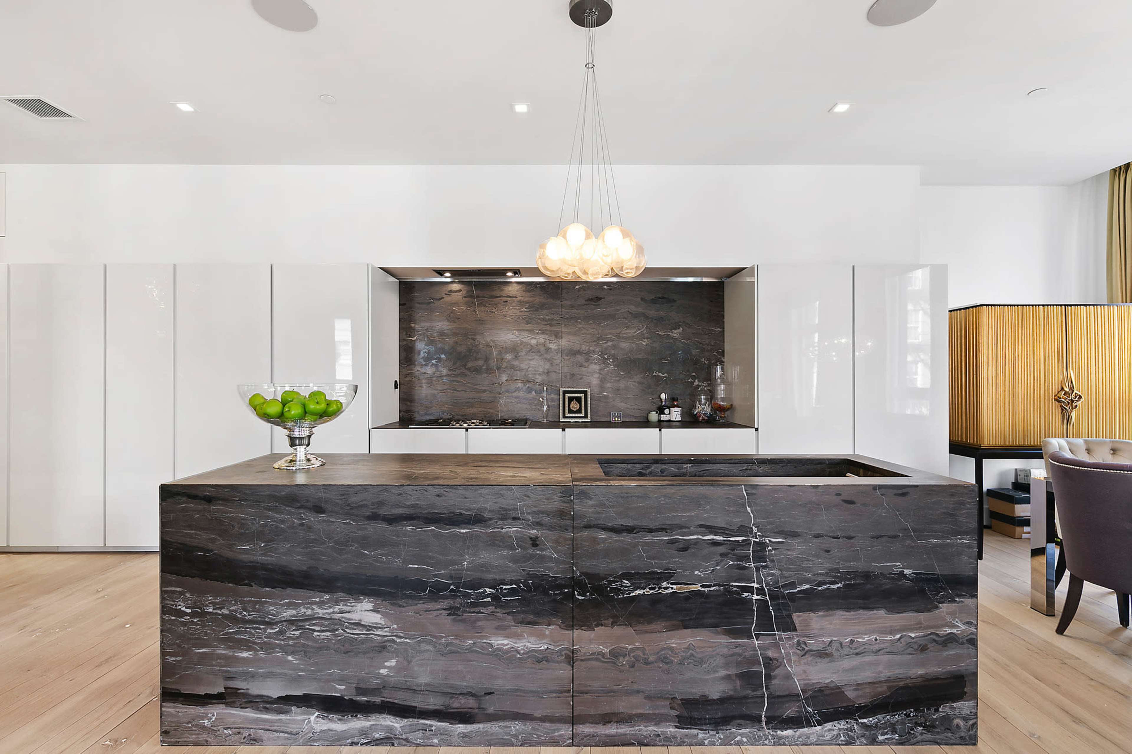 A modern kitchen features a large island with a dark stone countertop, sleek white cabinetry, and a statement light fixture above.