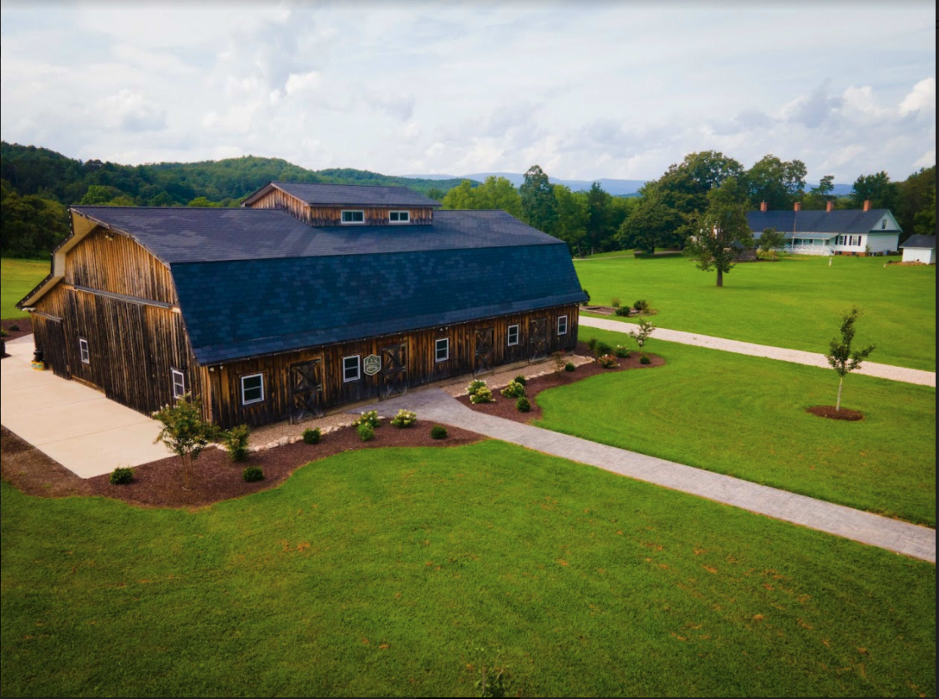 A large, rustic wooden building sits next to a well-maintained lawn and a paved pathway in a rural area surrounded by rolling hills.