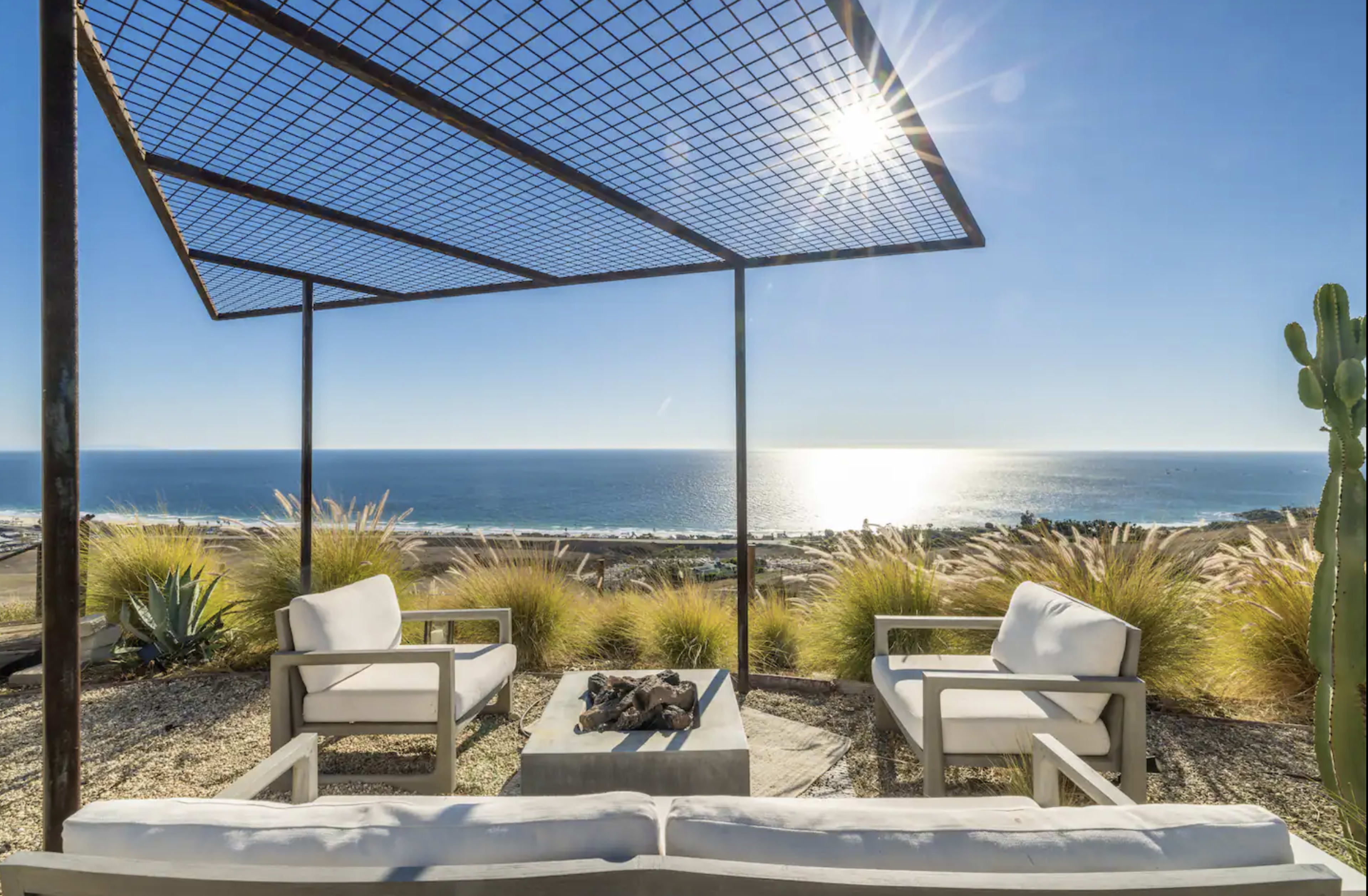 A modern outdoor seating area features white armchairs and a stone table around a fire pit, overlooking the ocean under a lattice canopy.