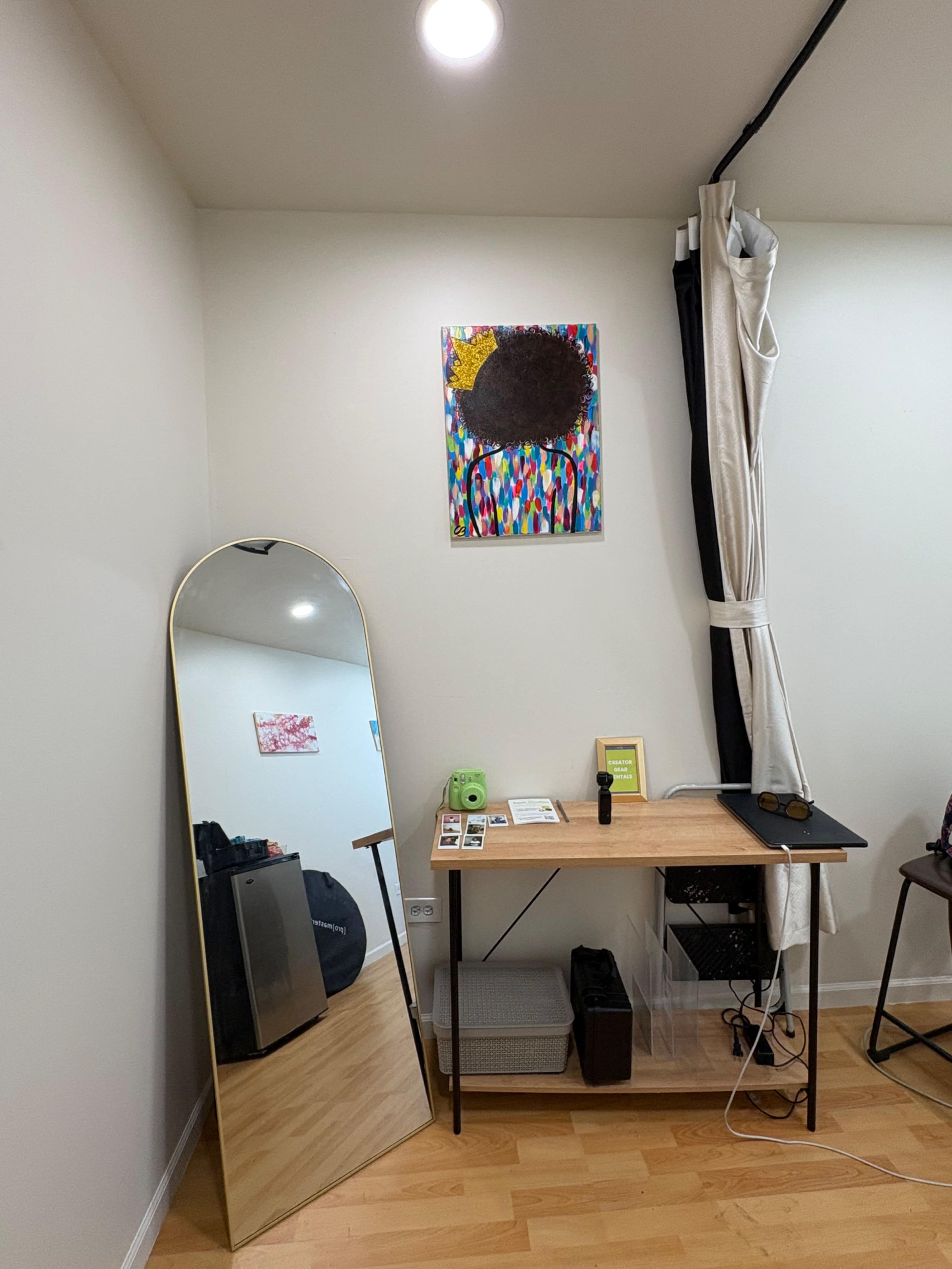 The image shows a minimalist room with a large mirror on the left, a wooden desk with various items on top, and a colorful artwork hanging on the wall above the desk.