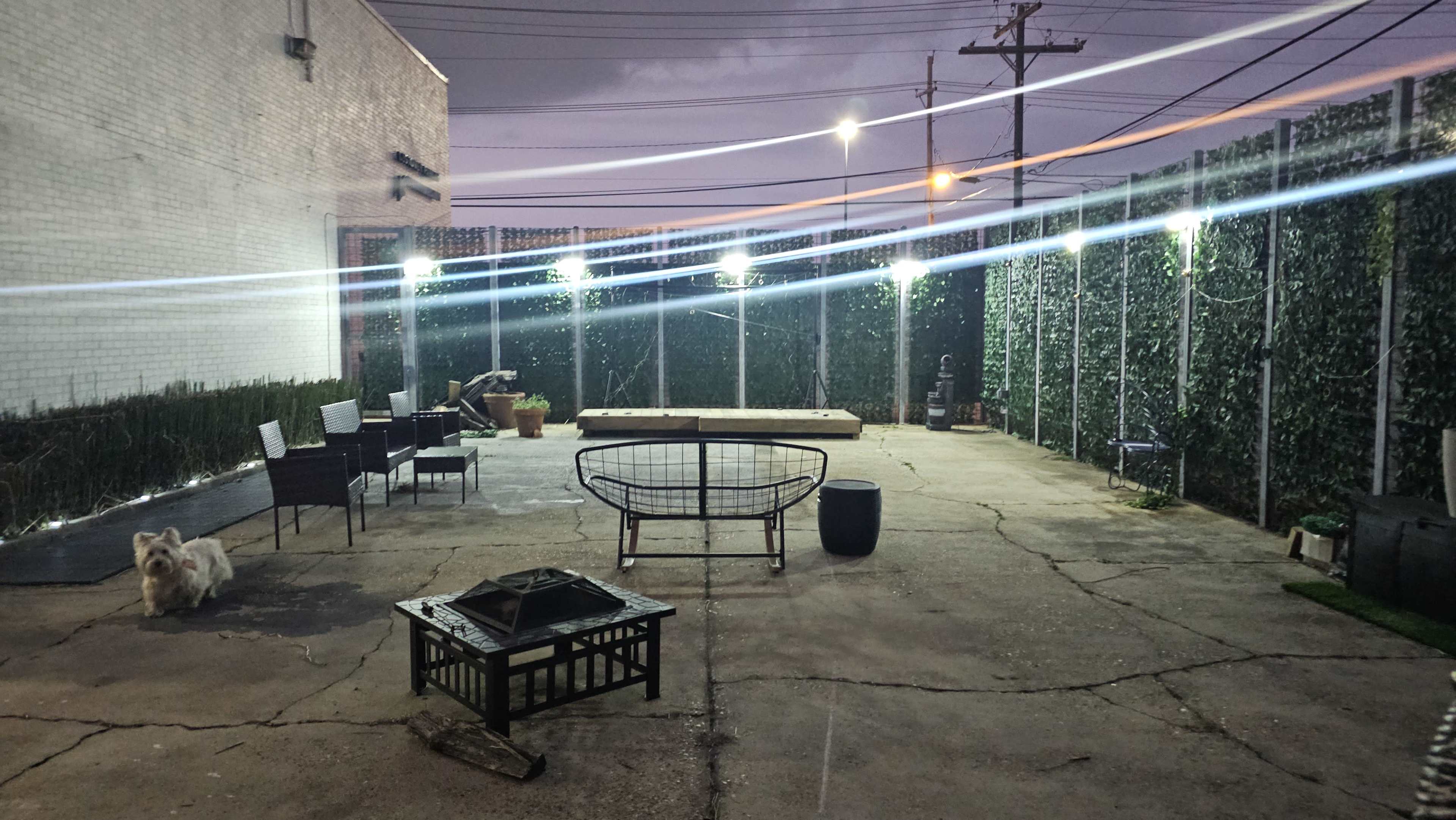 The image shows a dimly lit outdoor space with a concrete floor, various pieces of patio furniture, and a small dog on the left.