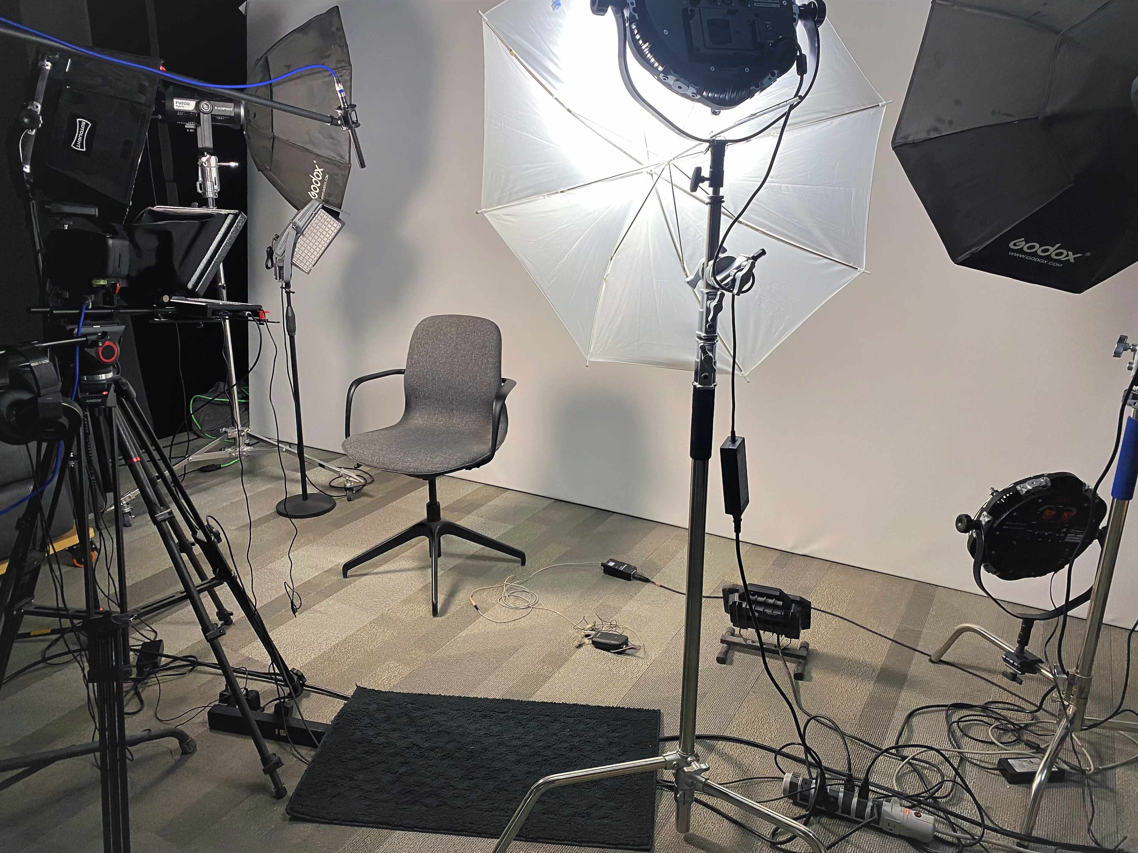 A filming setup with a gray chair under softbox lights, surrounded by camera equipment and a backdrop.