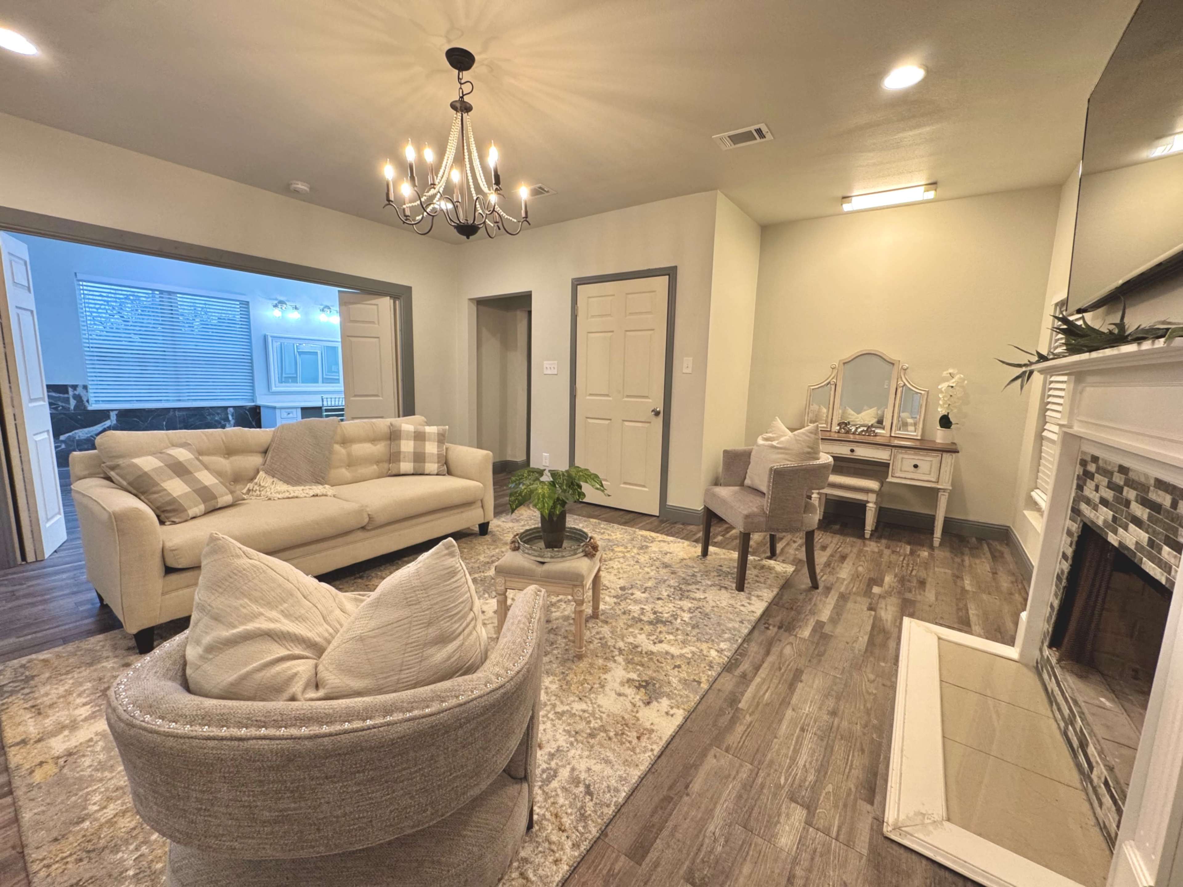 The image shows a living room with a beige sofa, two armchairs, a chandelier, and a small table, all arranged on a patterned rug, with a fireplace visible on one side.