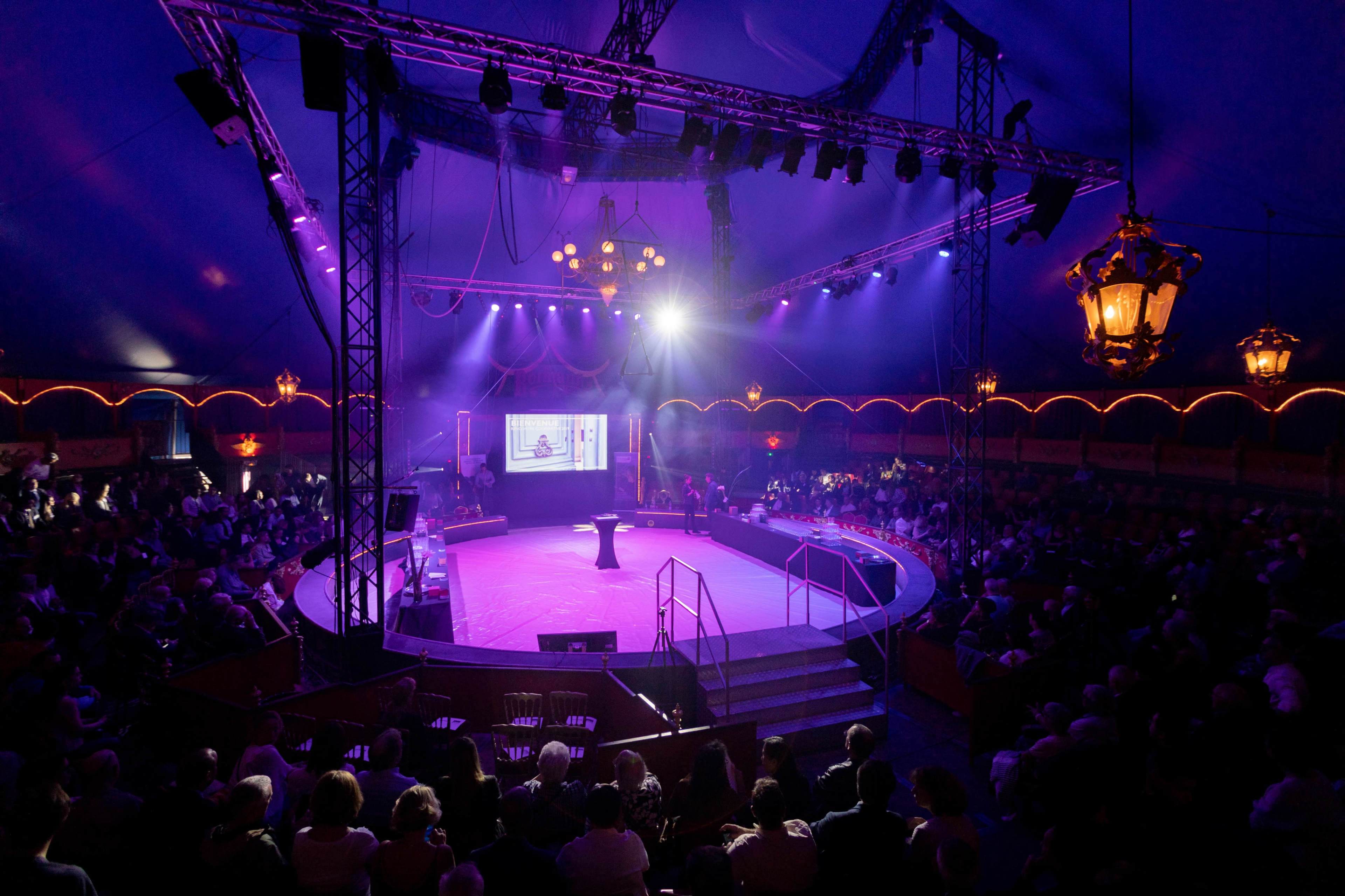 A crowd gathers under a large circus tent with purple lighting, centered around a circular stage.
