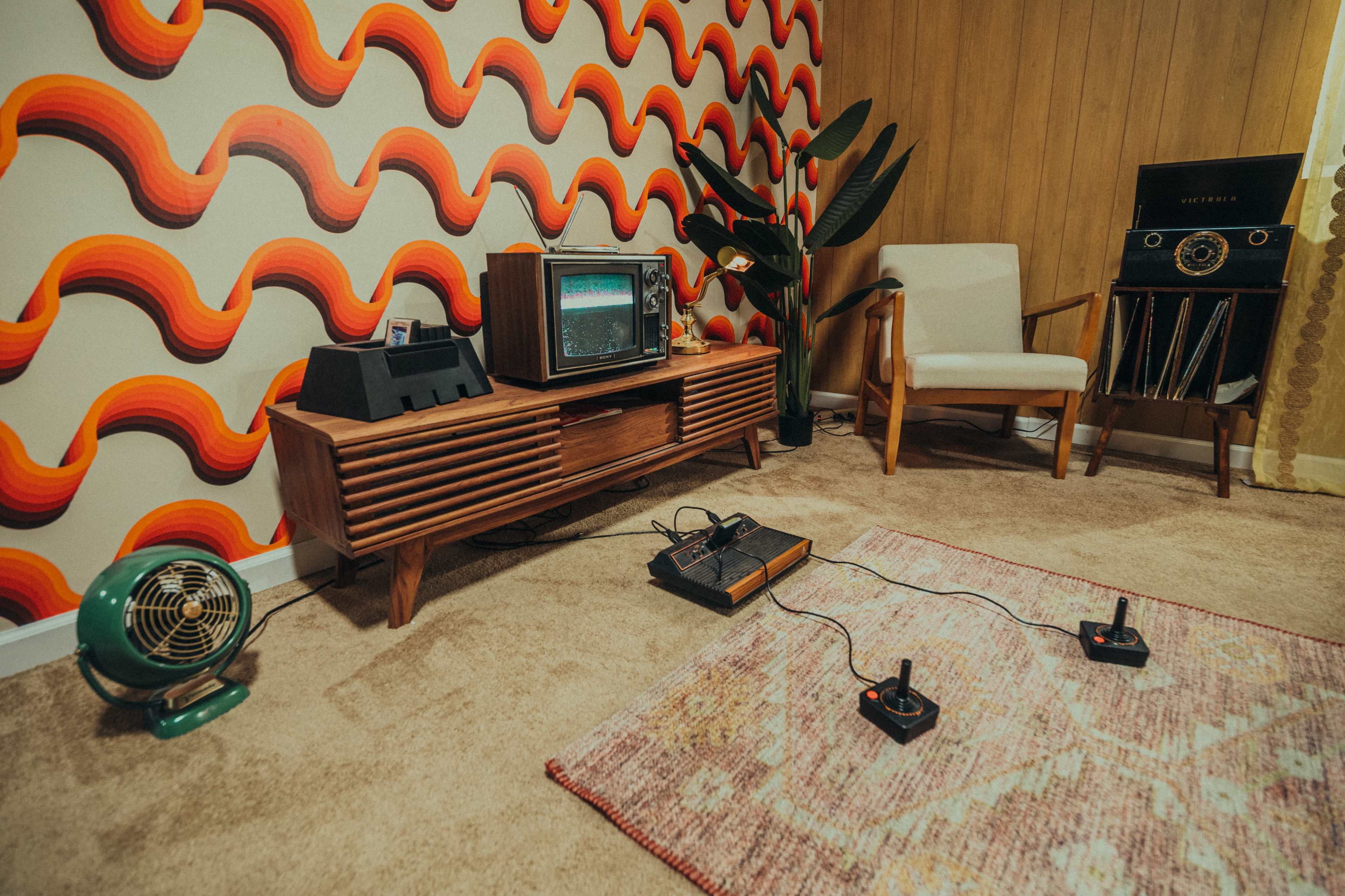 A retro living room with a vintage television, a wooden TV stand, an Atari console connected to two controllers, a green fan, and a patterned wallpaper featuring wavy orange and yellow shapes.