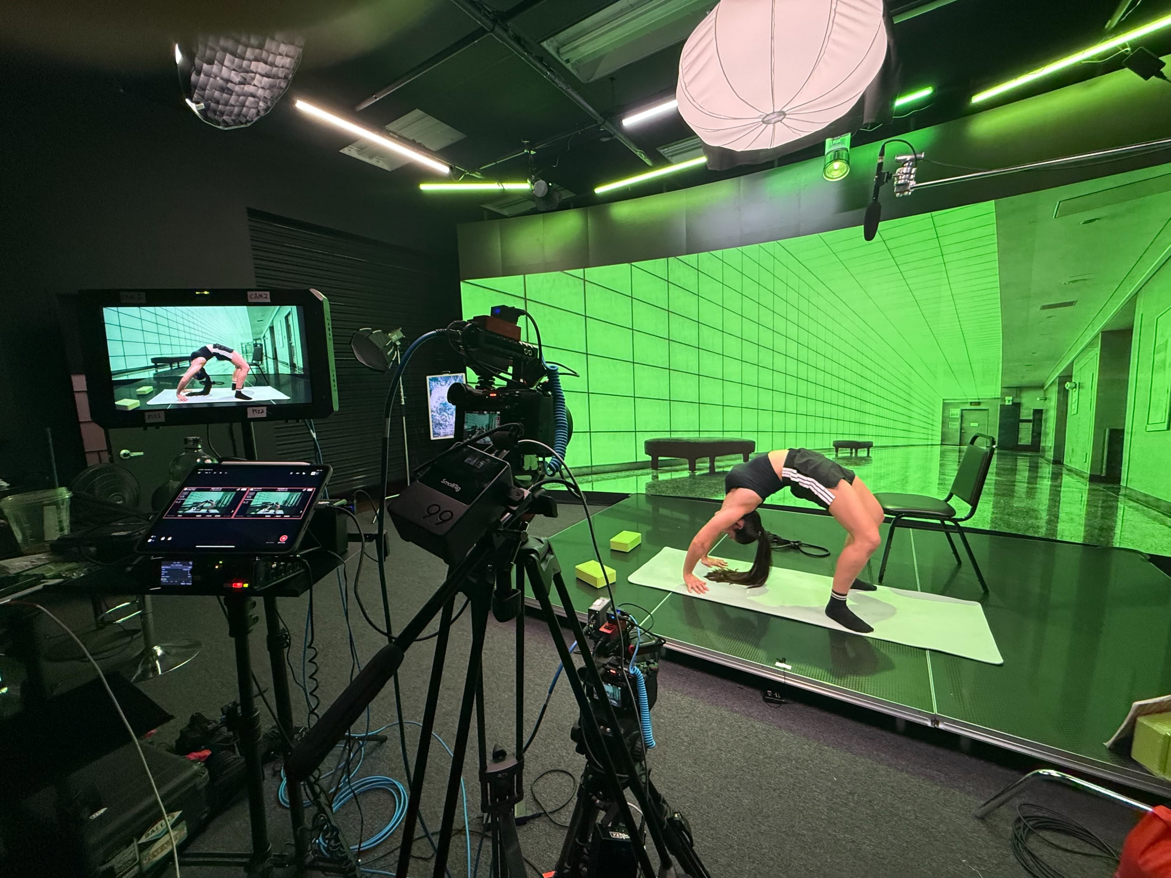 A person is performing a yoga stretch on a mat in a studio with green lighting and a monitor displaying their performance.