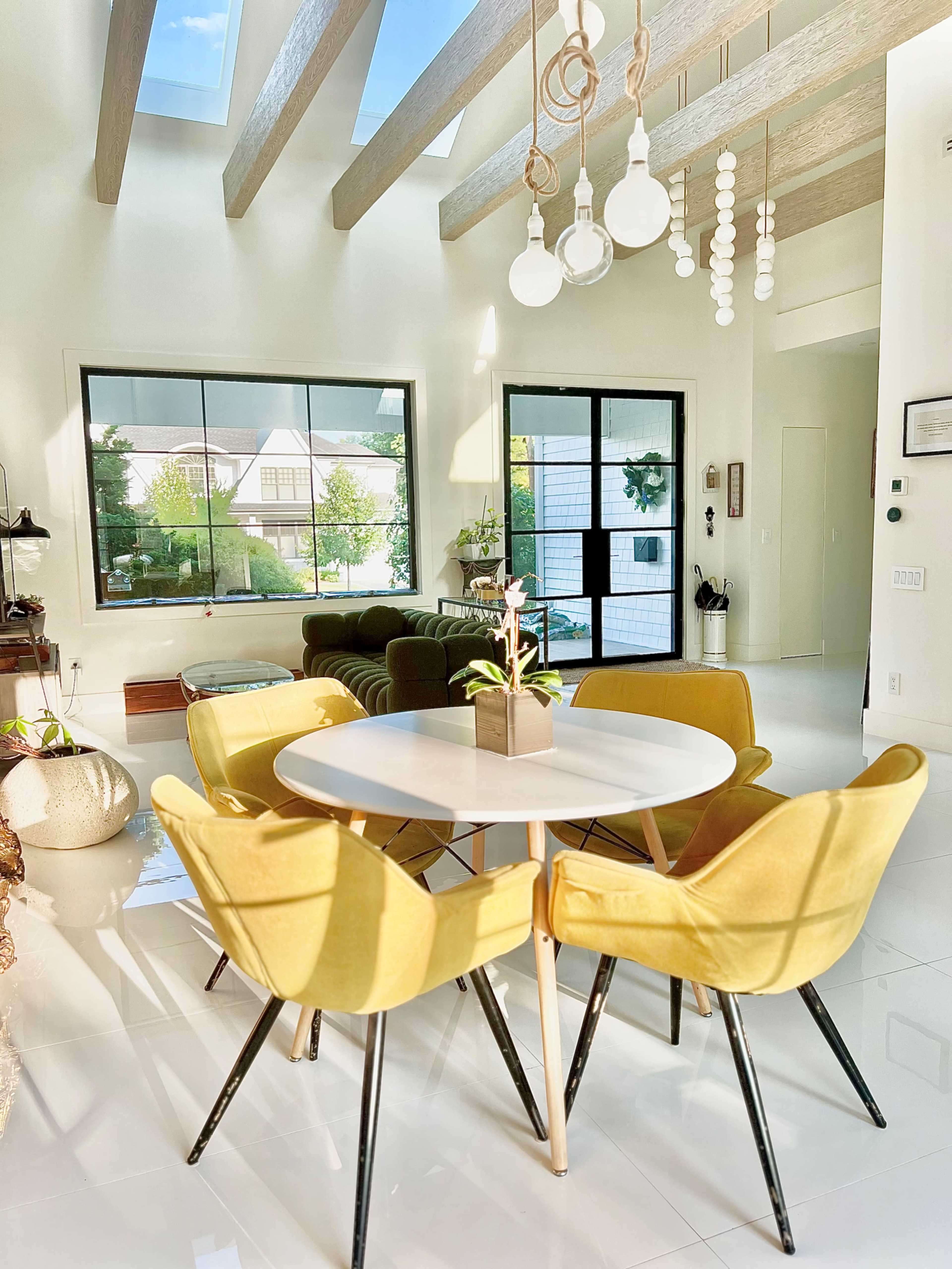 A modern dining area features a round white table surrounded by yellow chairs, with large windows allowing natural light to fill the space.