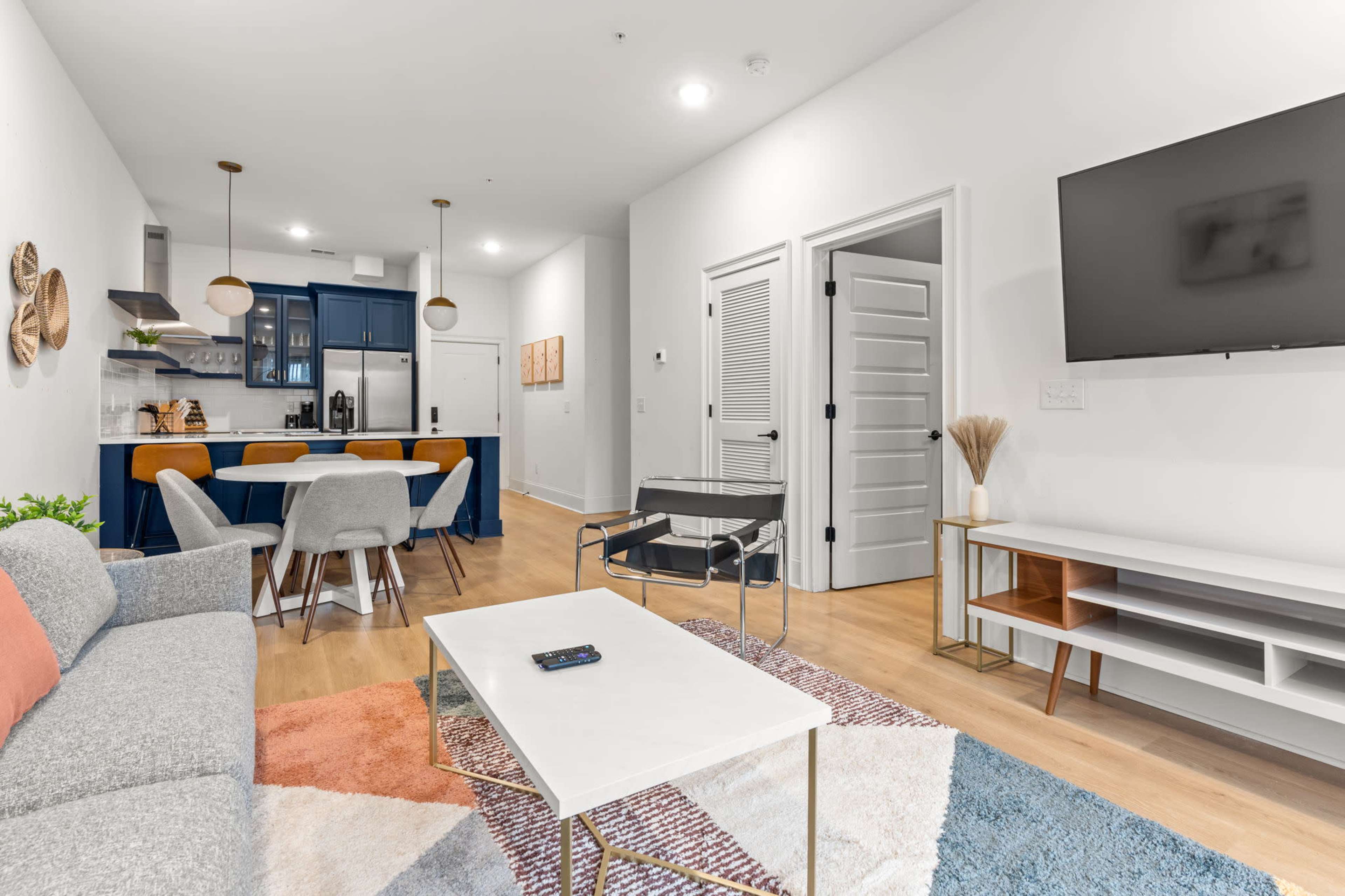 The image shows a modern living room with a gray sofa, a coffee table on a colorful rug, and a kitchen area visible in the background.