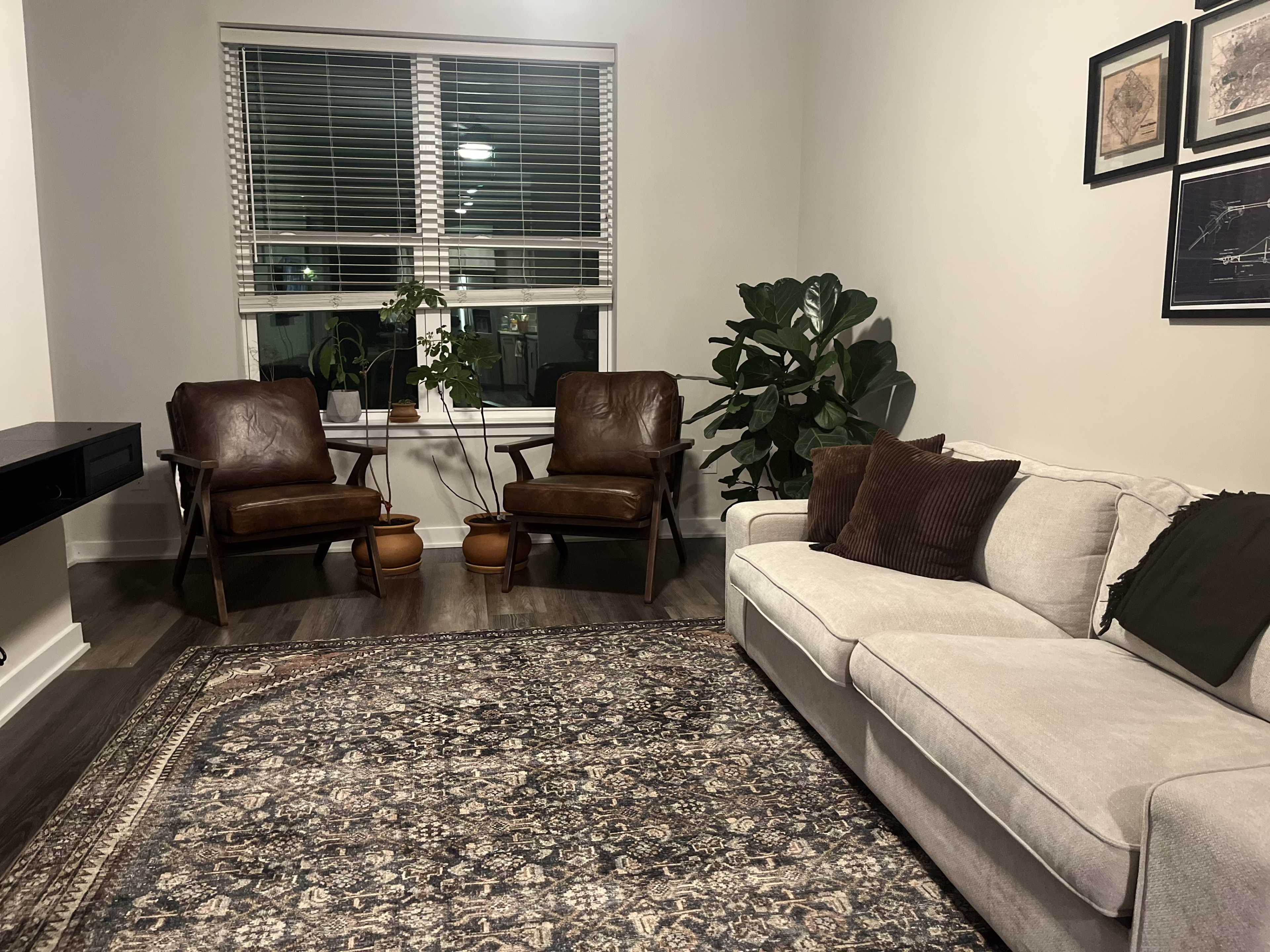The scene features a cozy living room with two brown armchairs and a light gray sofa, positioned around a patterned area rug beneath a window with blinds.