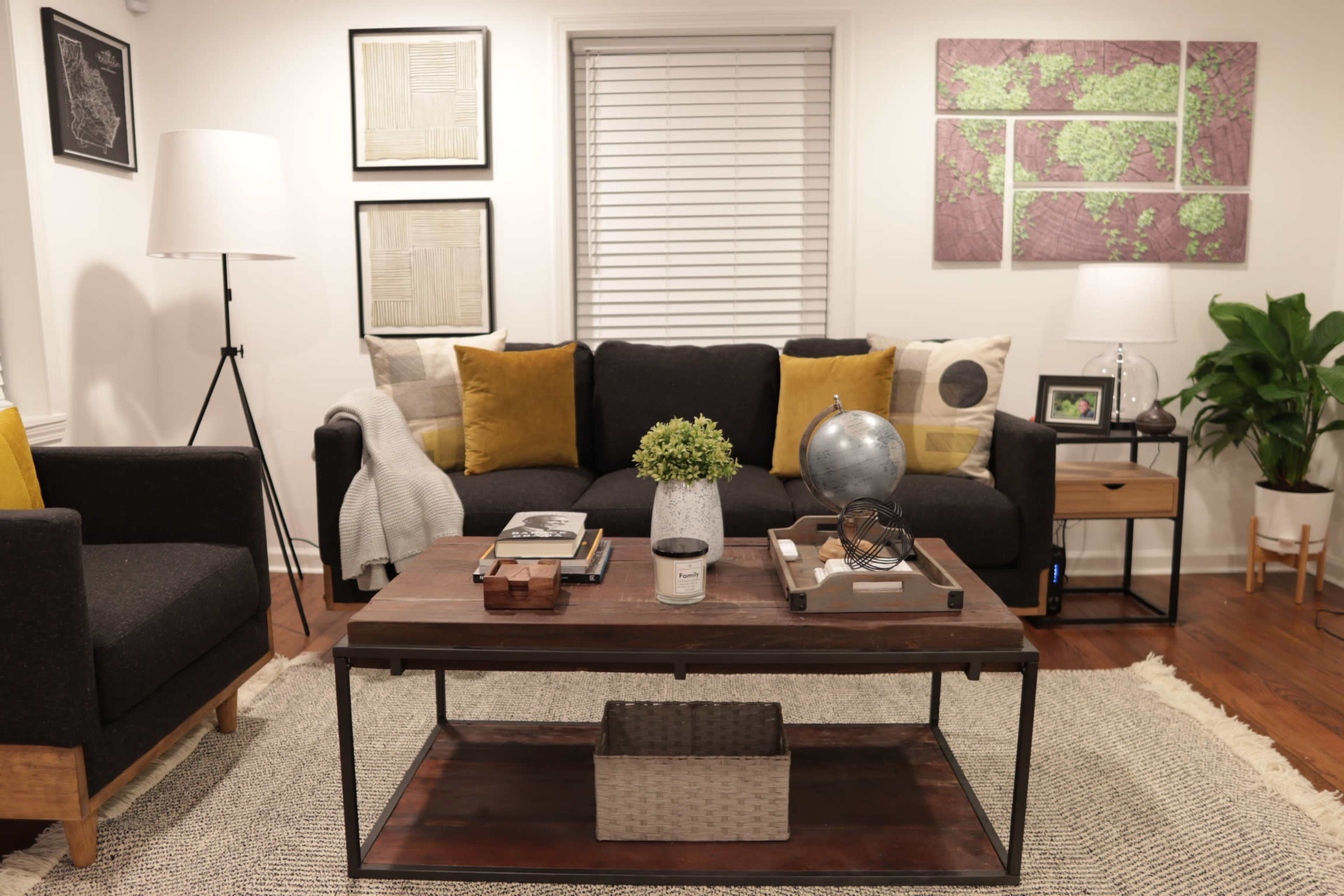 A living room with a dark sofa, a wooden coffee table, framed artwork on the walls, and a potted plant in the corner.