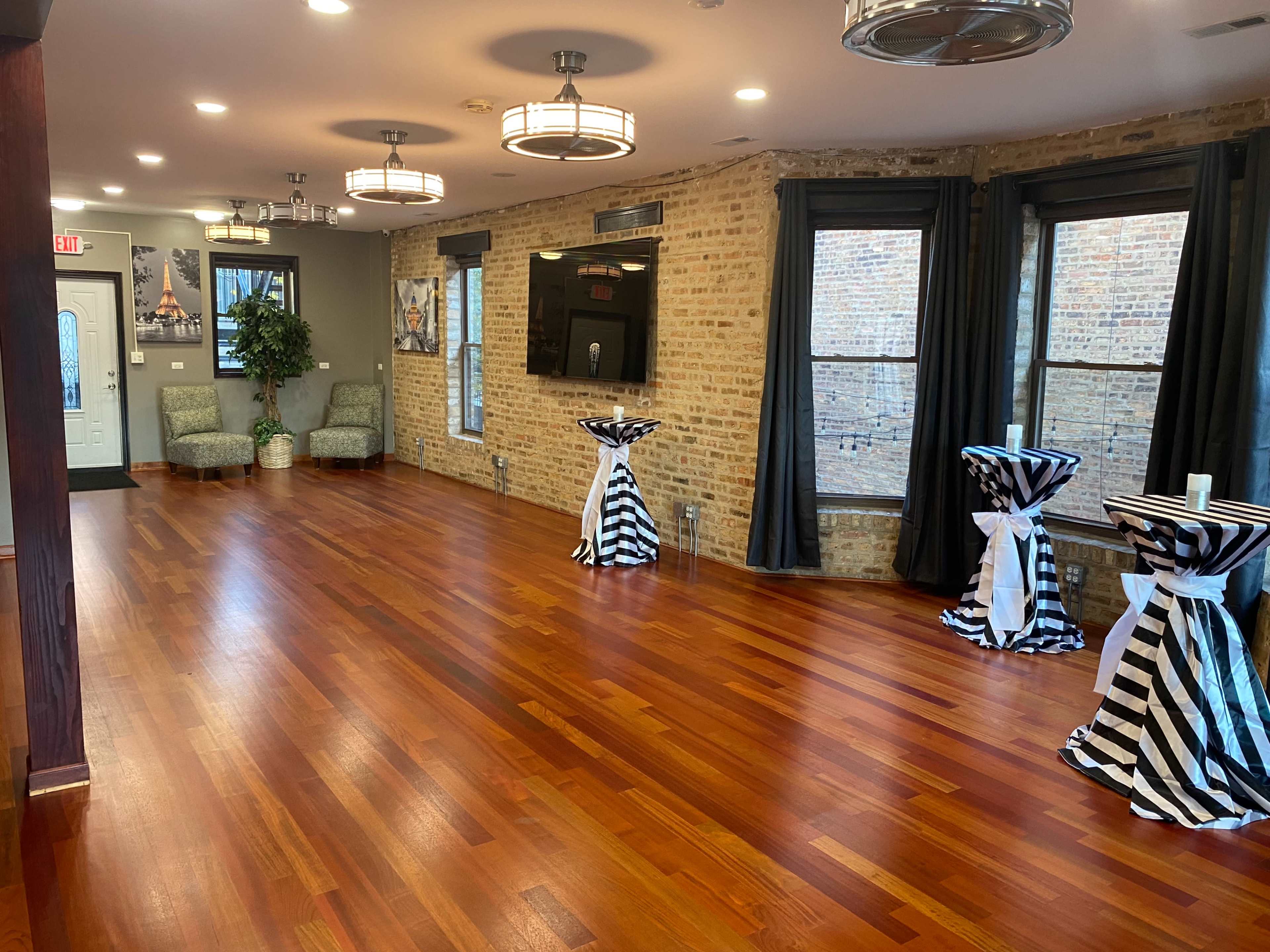 A spacious interior room with polished wooden floors, decorative papered walls, and a few striped cocktail tables, illuminated by modern light fixtures.