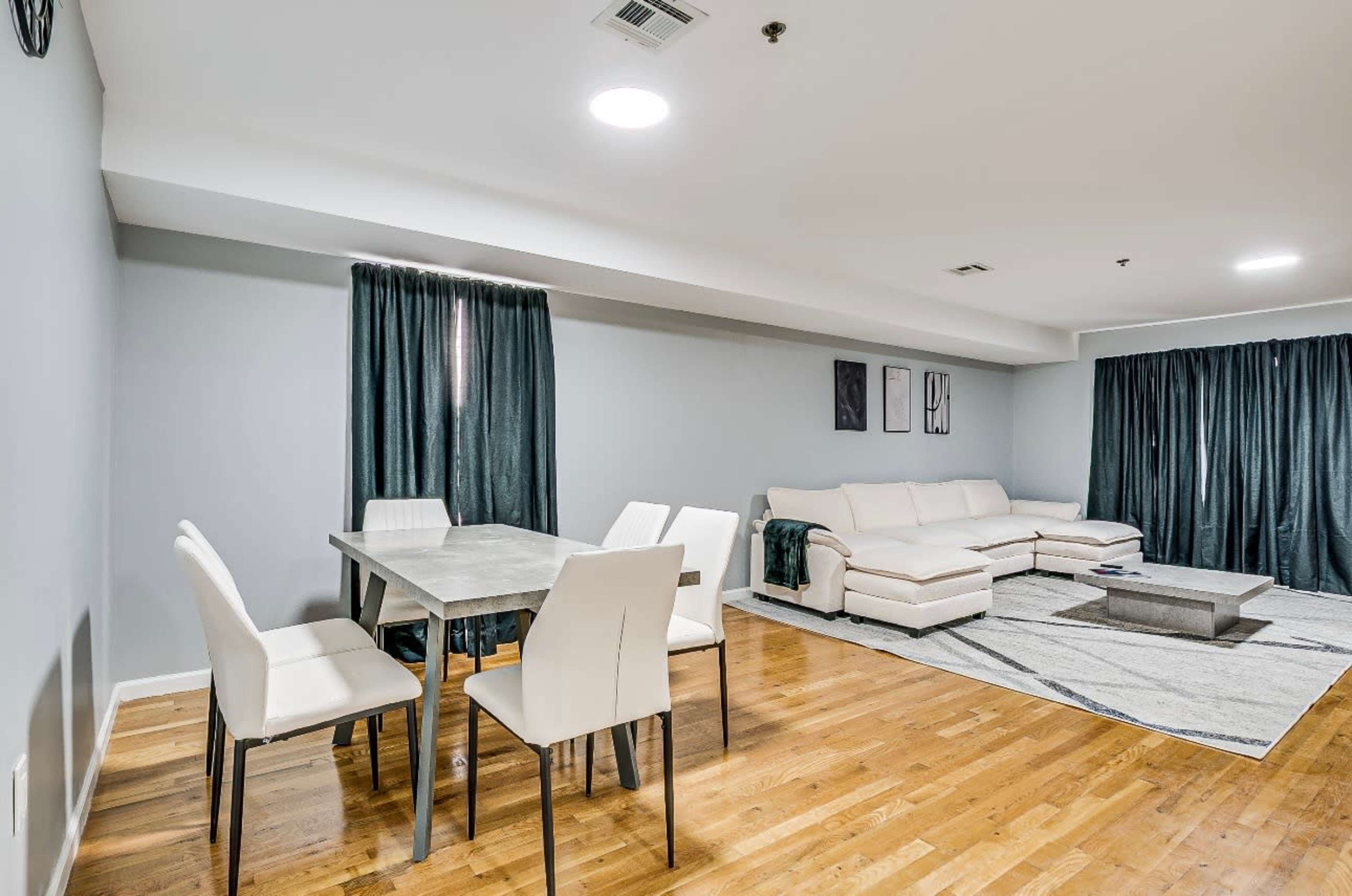 The image shows a modern living and dining area featuring a white sectional sofa, a rectangular dining table with four chairs, and dark curtains covering the windows.