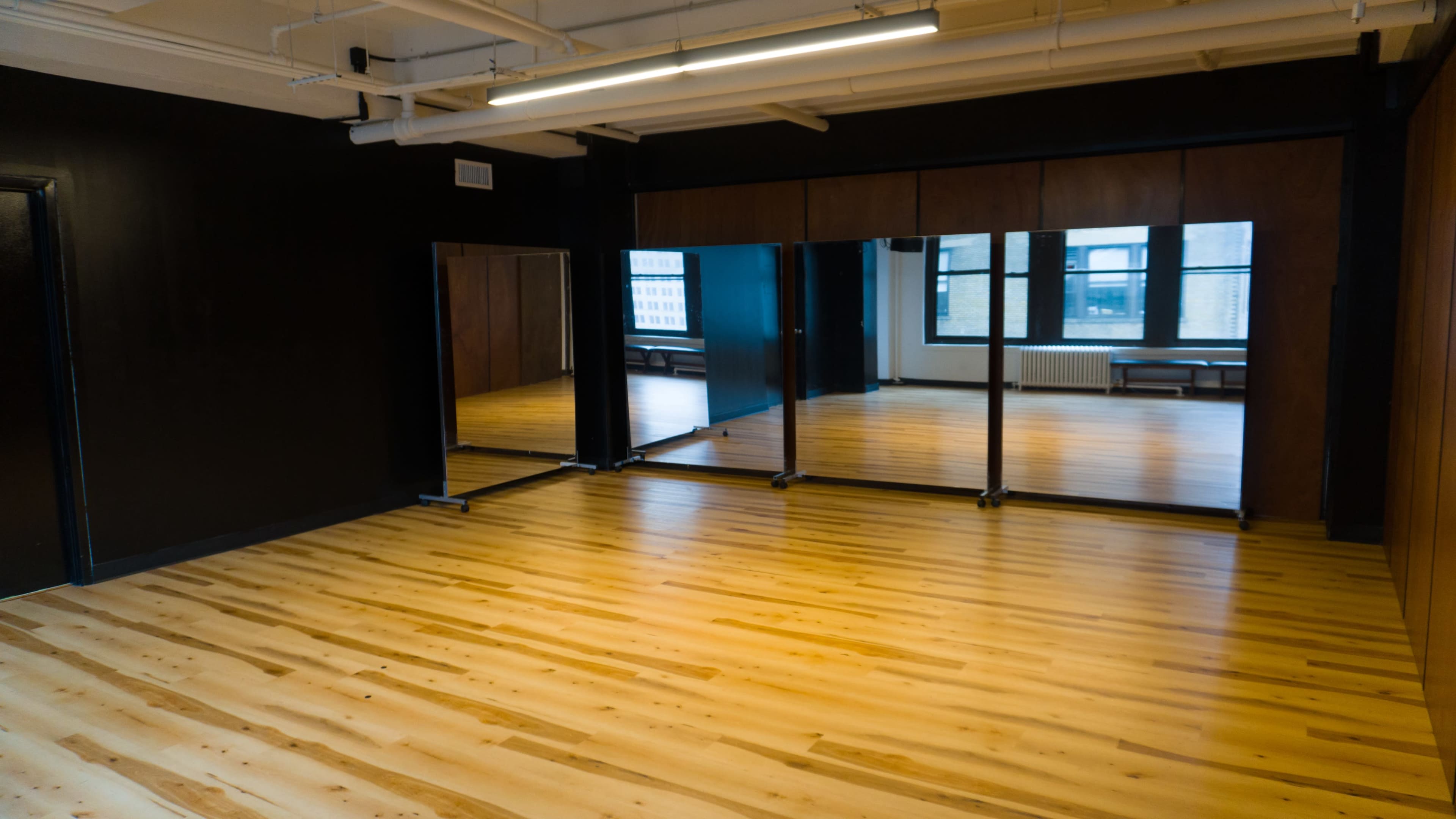 The image shows a spacious room with wooden flooring, large mirrors on one wall, and windows that provide natural light.