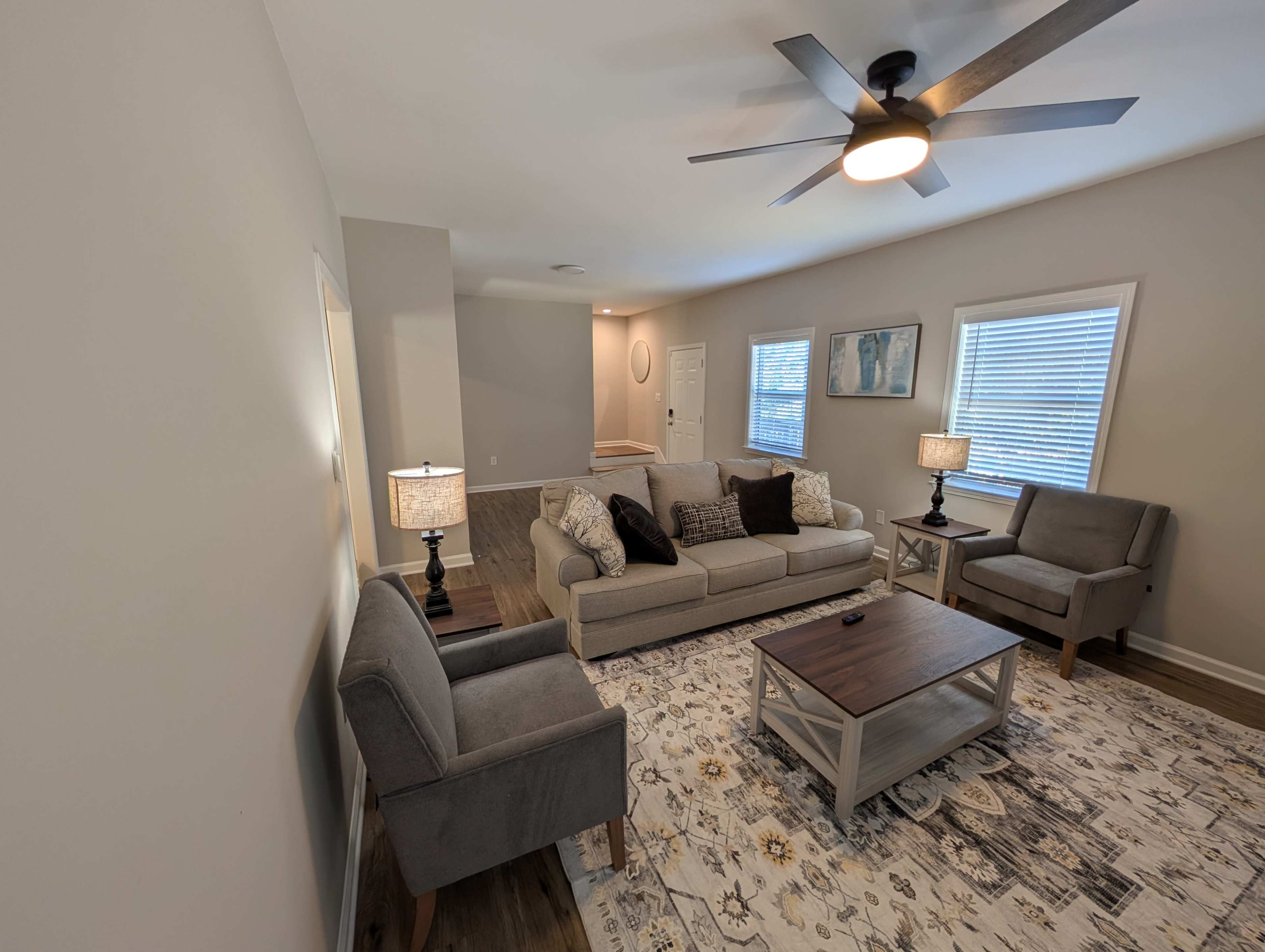 The image shows a living room with a light gray sofa, two armchairs, a coffee table, and lamps, all arranged around a patterned rug and illuminated by a ceiling fan.