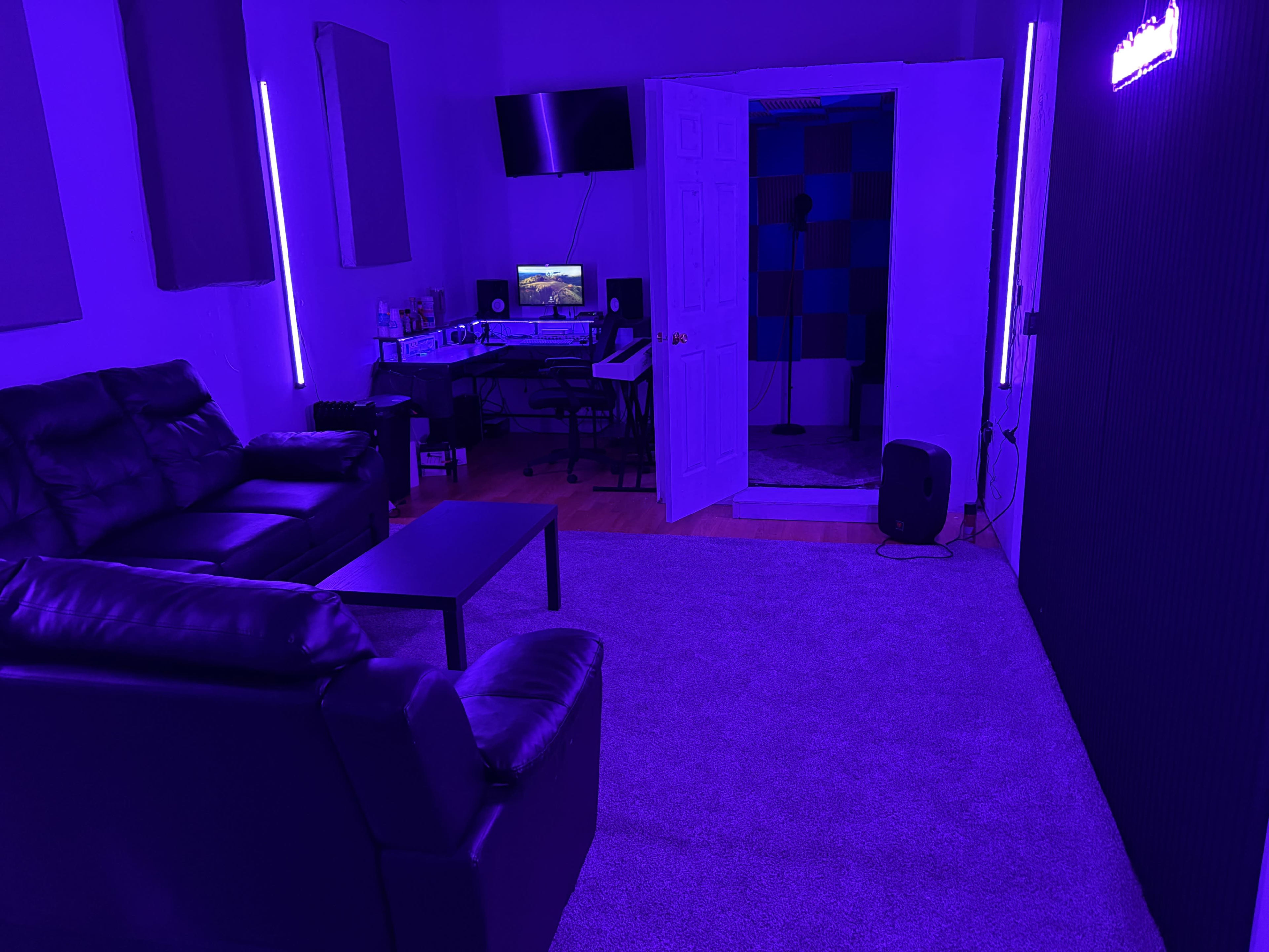 The image shows a room with purple lighting, featuring a black leather sofa, a small coffee table, a work desk with a computer, and a soundproof booth in an open doorway.