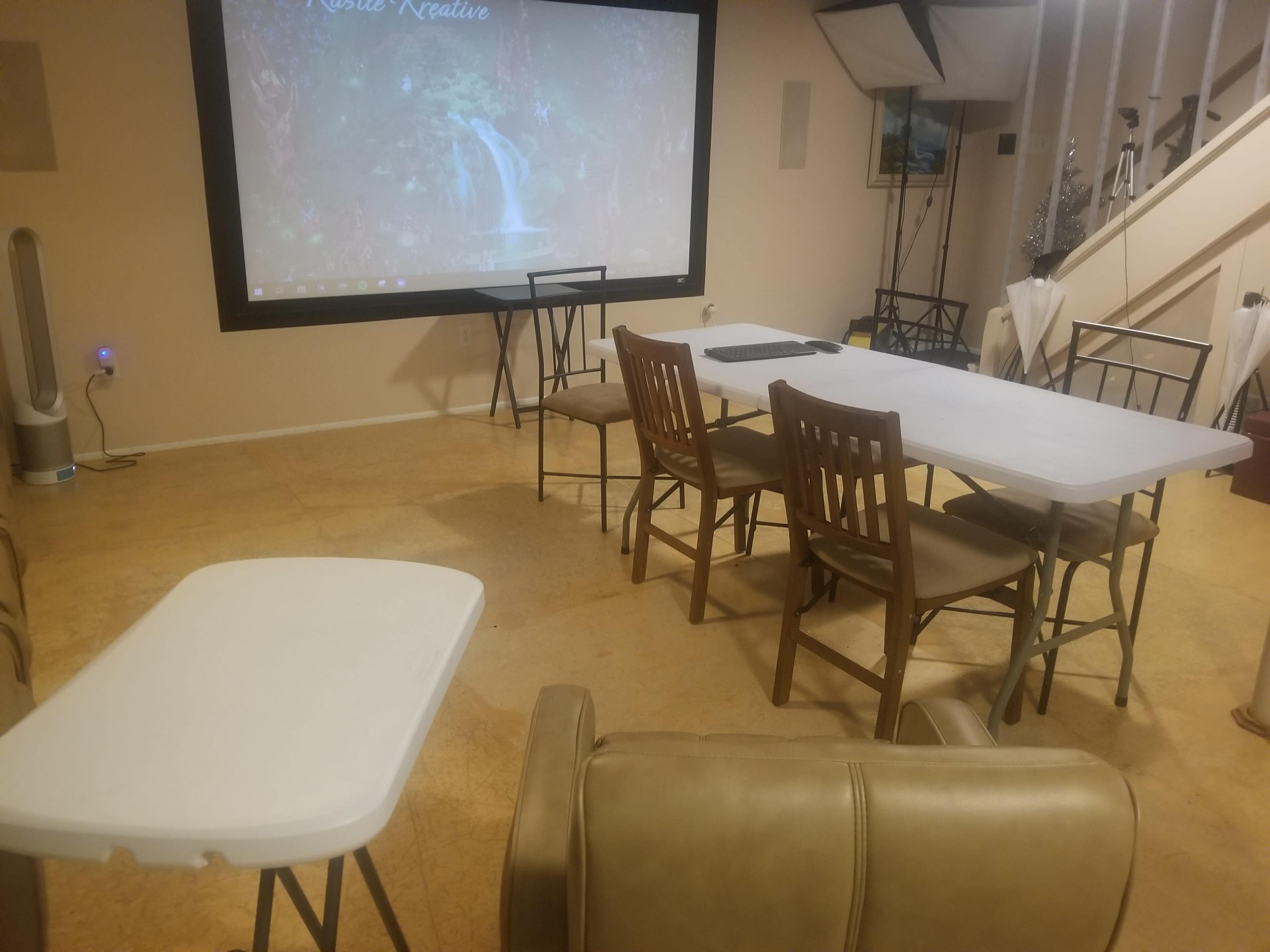 A small indoor room features a rectangular table surrounded by chairs, with a large screen displaying a nature scene on one wall.