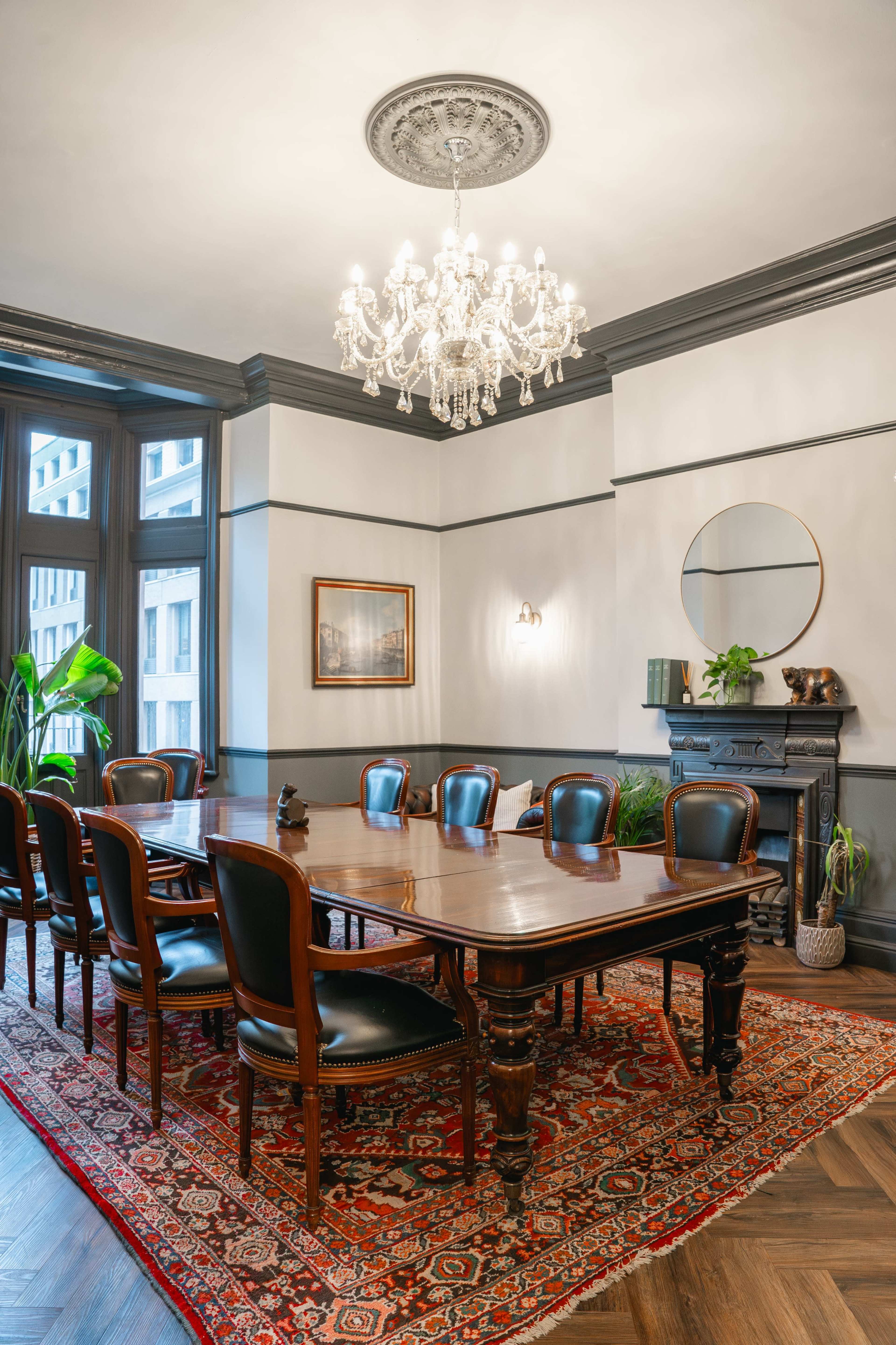 A large, elegant conference room features a polished wooden table surrounded by leather chairs, with a chandelier above and plants placed around the space.