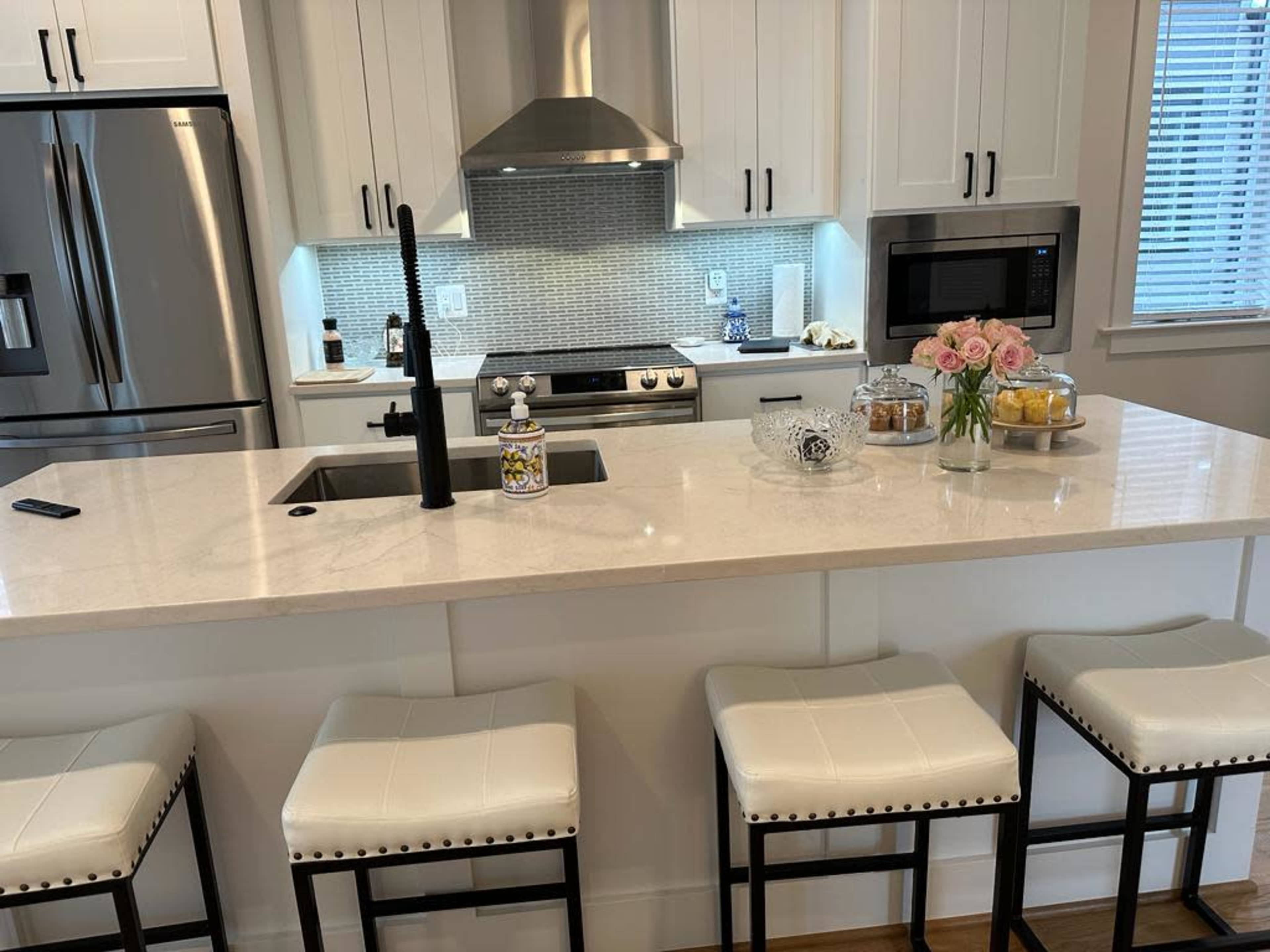 A modern kitchen with a central island, featuring a sink, bar stools, and countertop decor including flowers and a glass bowl.