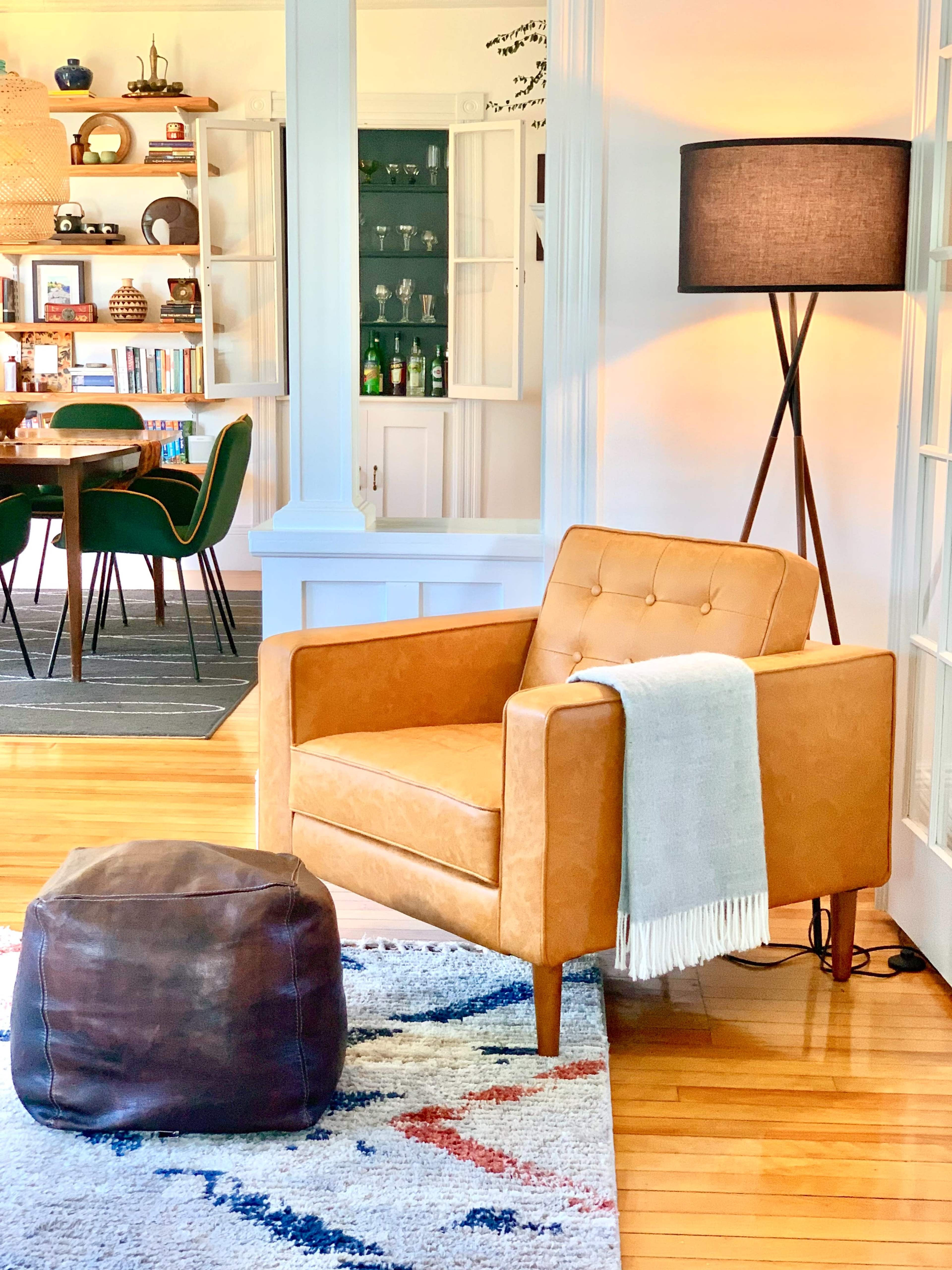 A tan armchair with a throw blanket and a brown ottoman on a patterned rug, positioned near a tall floor lamp in a bright living space.