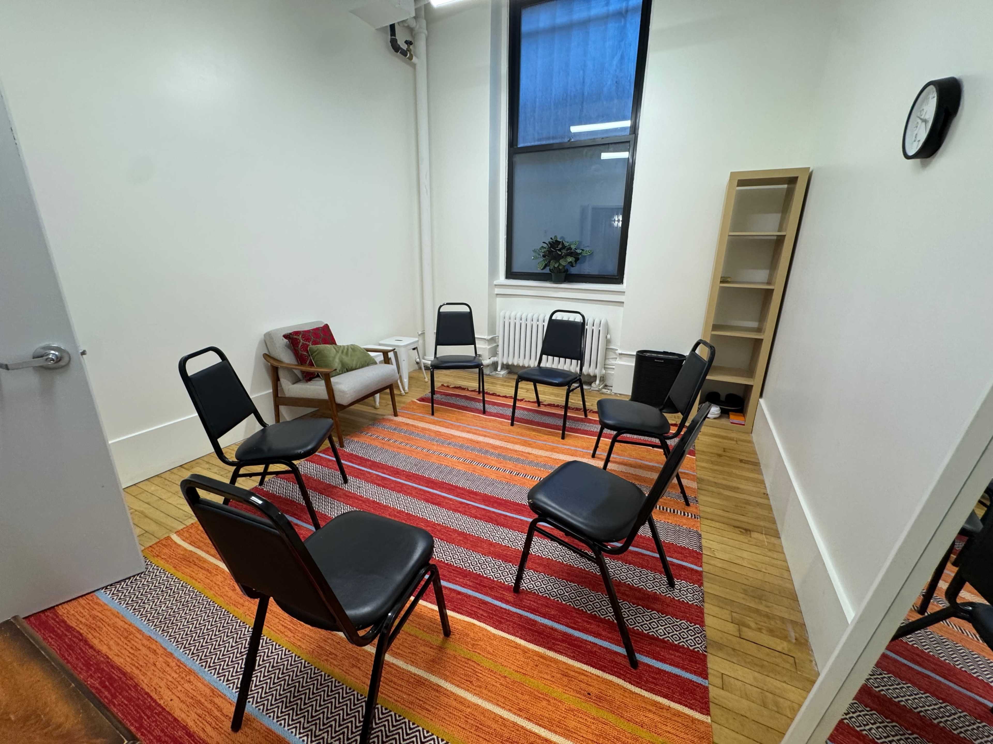 A small, empty room features a circular arrangement of black chairs on a colorful rug, with a chair and a plant in one corner, and a shelving unit against the wall.