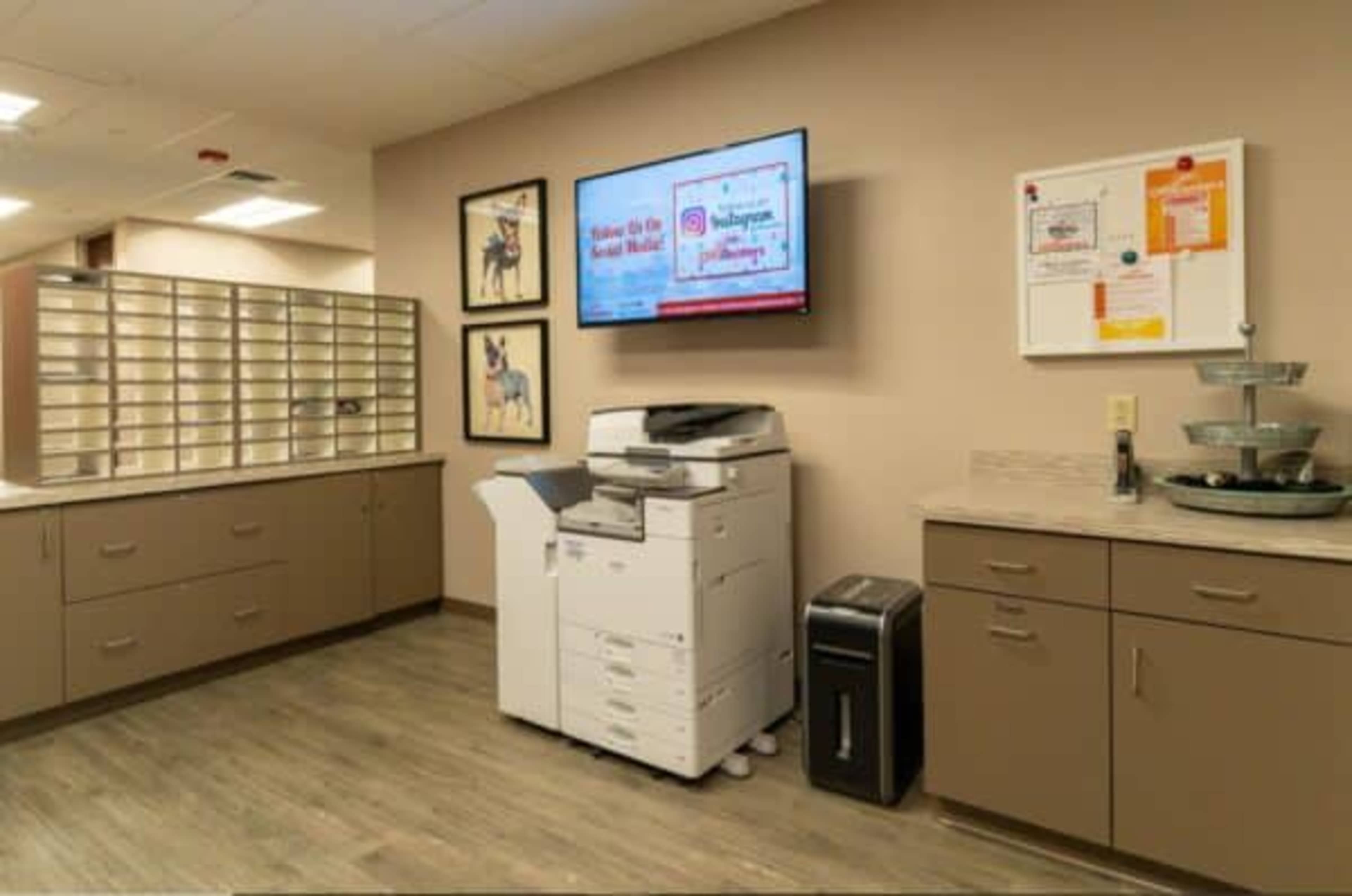 The image shows an office space with a printer, cabinets, and a large monitor displaying information on the wall.