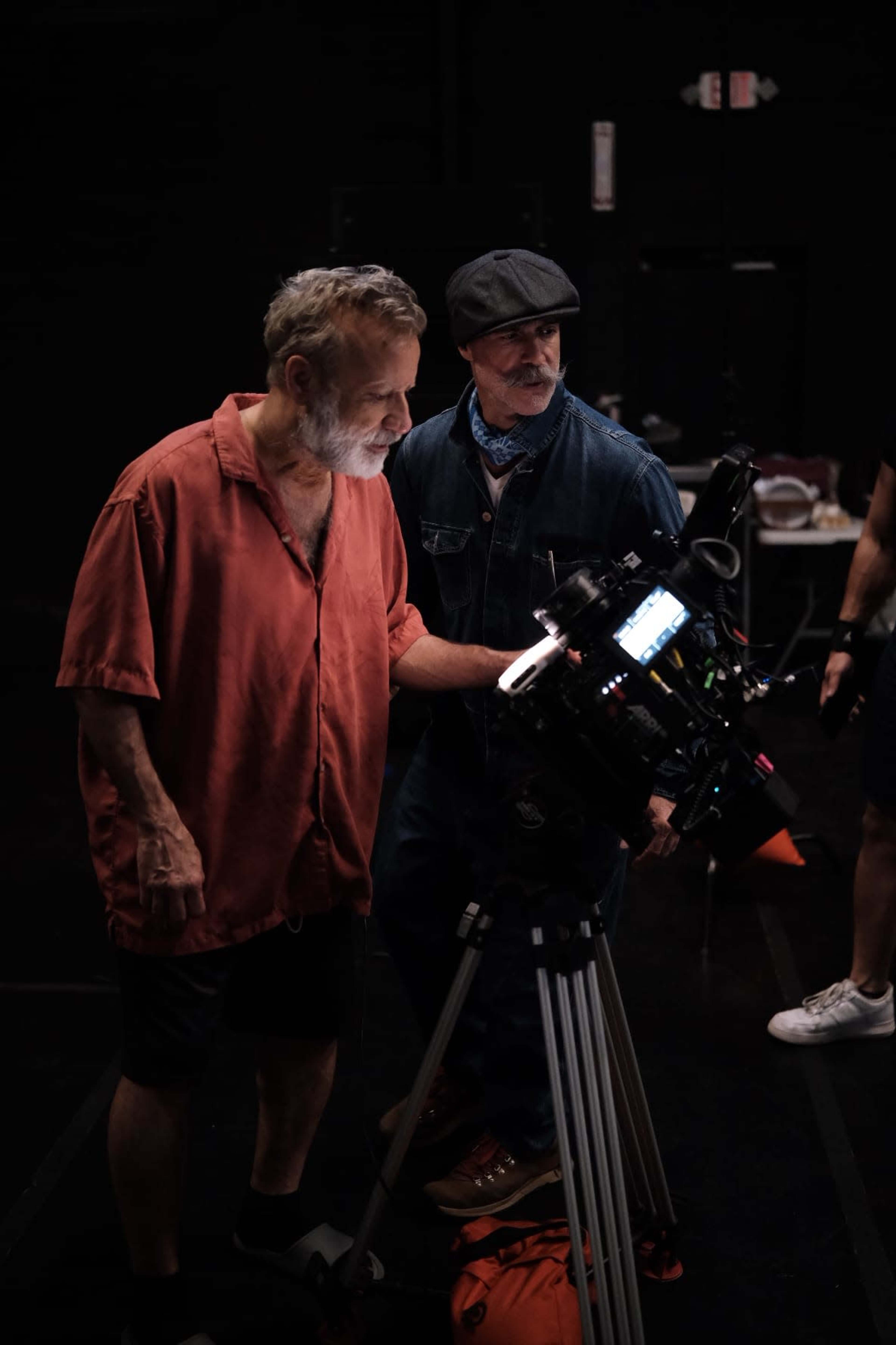 Two men are observing and adjusting a camera setup on a tripod in a dimly lit studio.