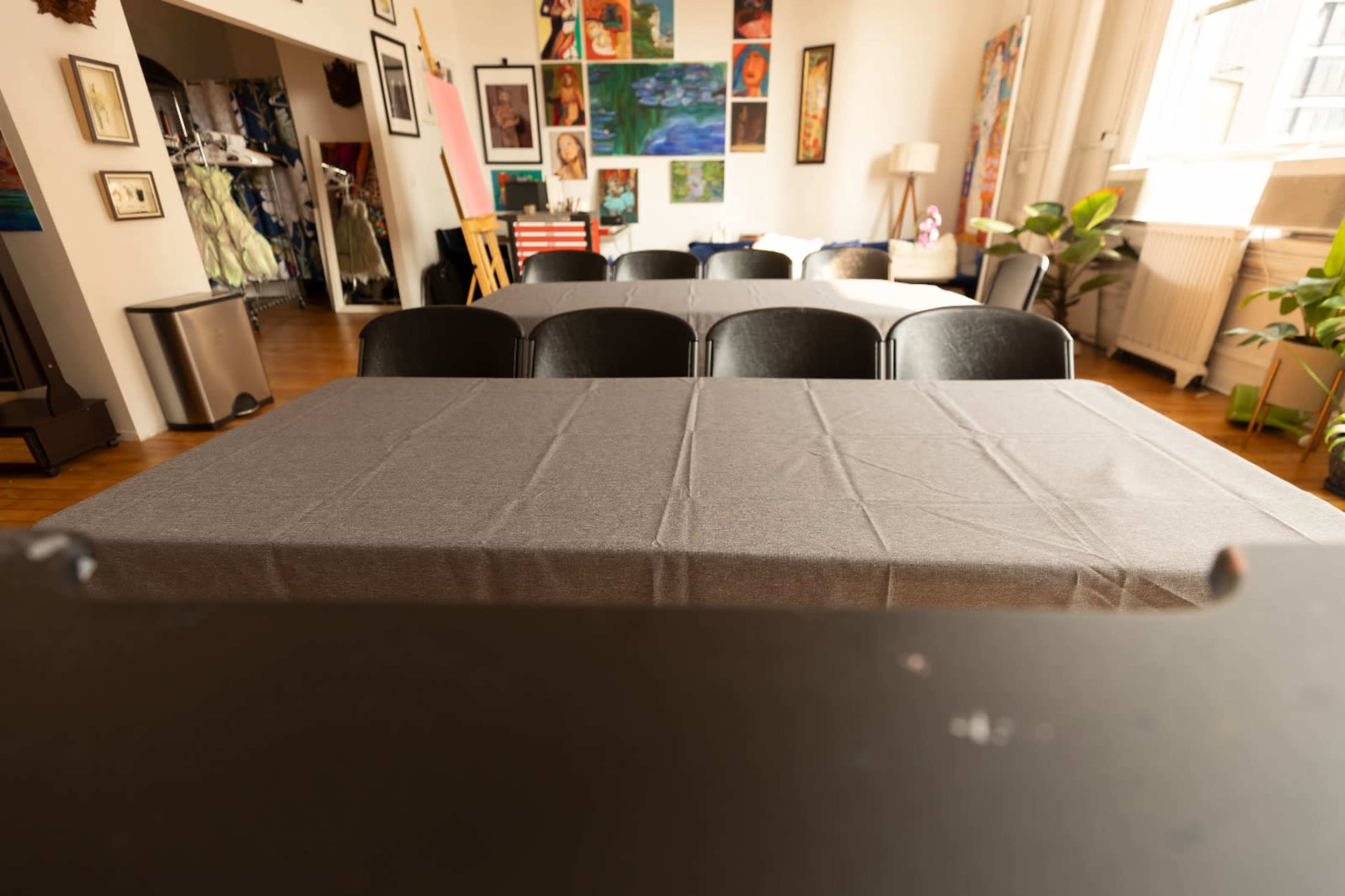 A large, empty room features a long table covered with a gray tablecloth, surrounded by black chairs, with colorful artwork on the walls in the background.