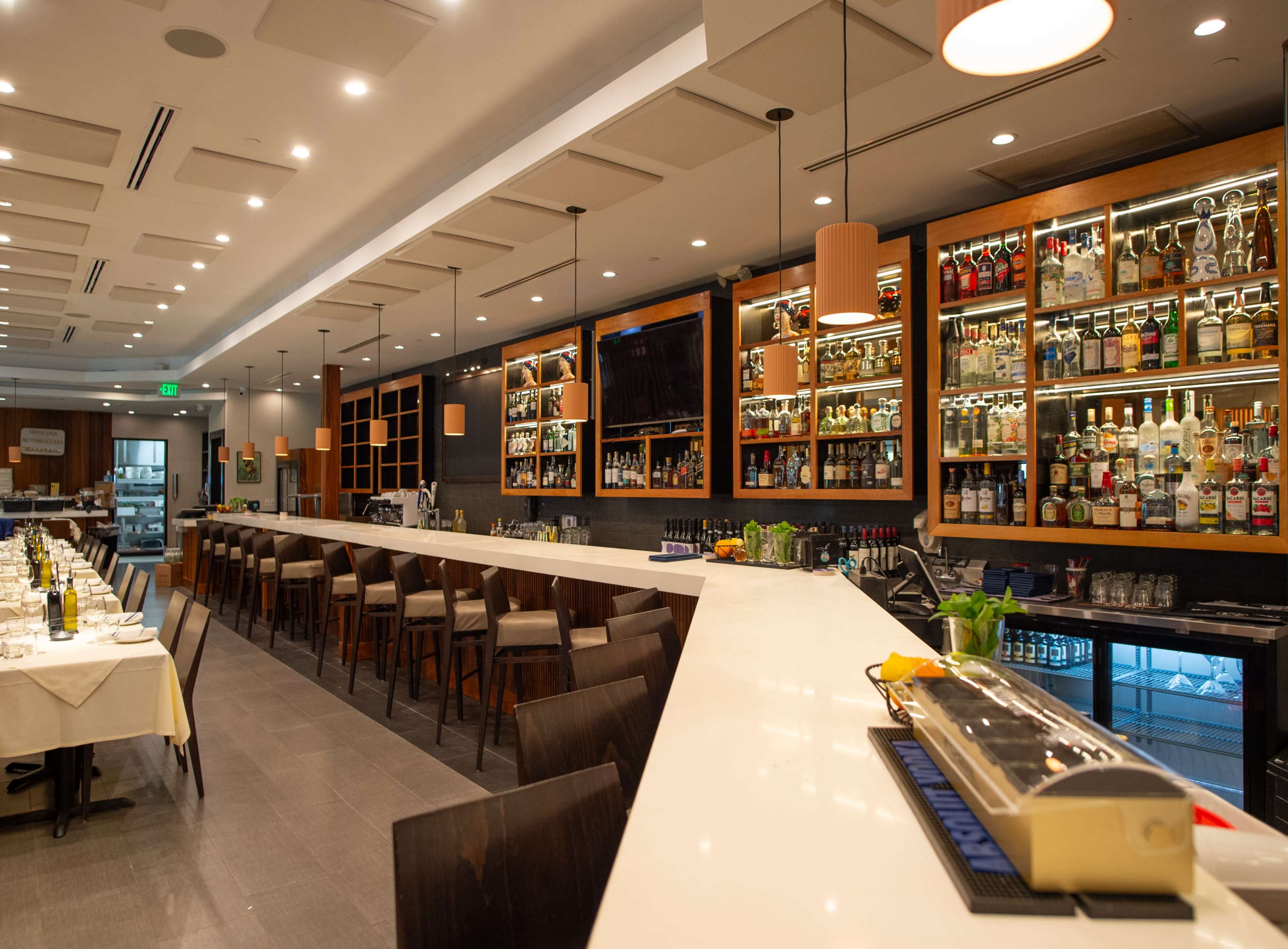 A modern bar area features a long white counter, wooden stools, and shelves lined with various spirits and liquors.